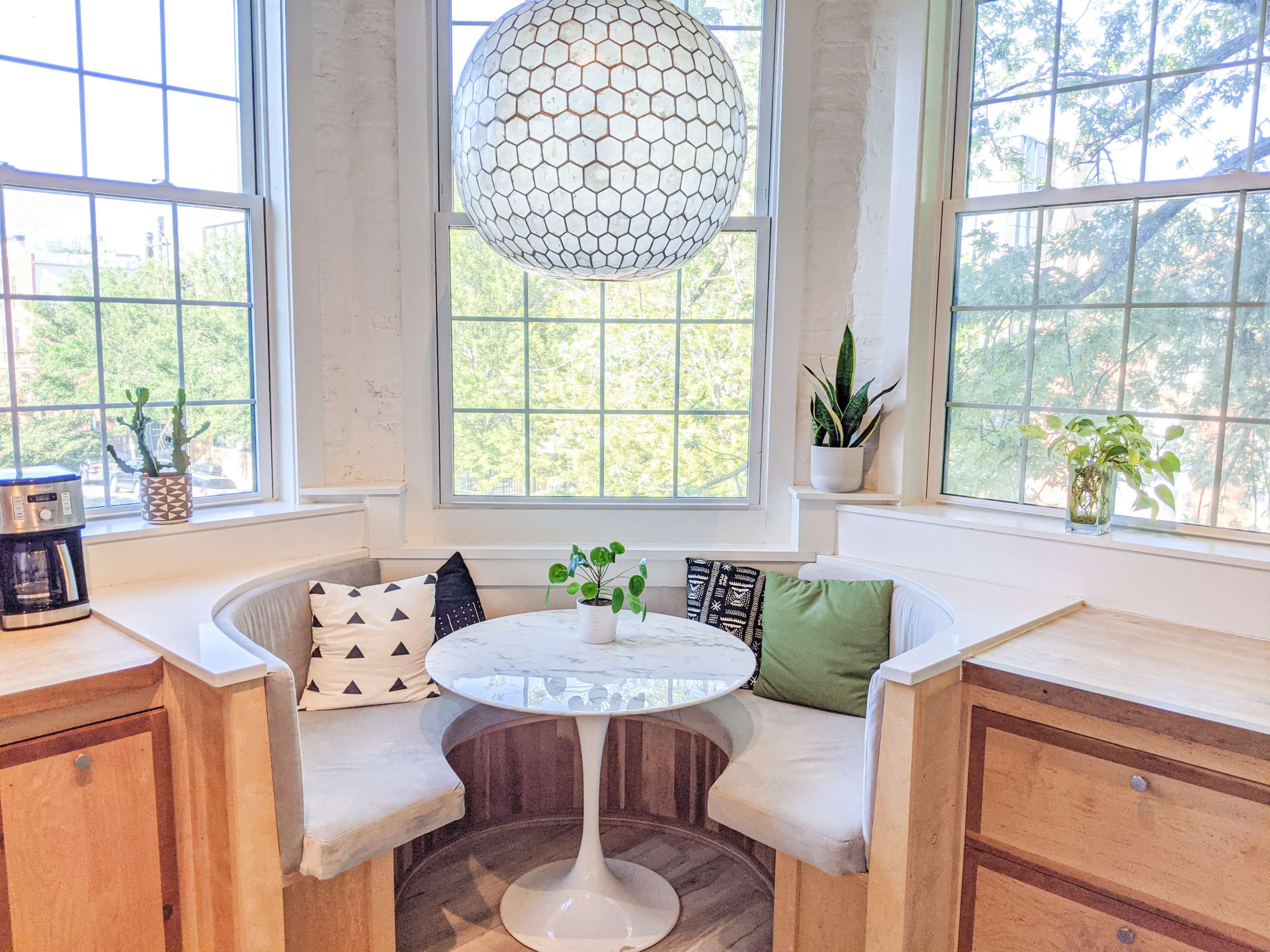 A cozy breakfast nook features a round table surrounded by curved seating, with large windows letting in natural light and plants on display.