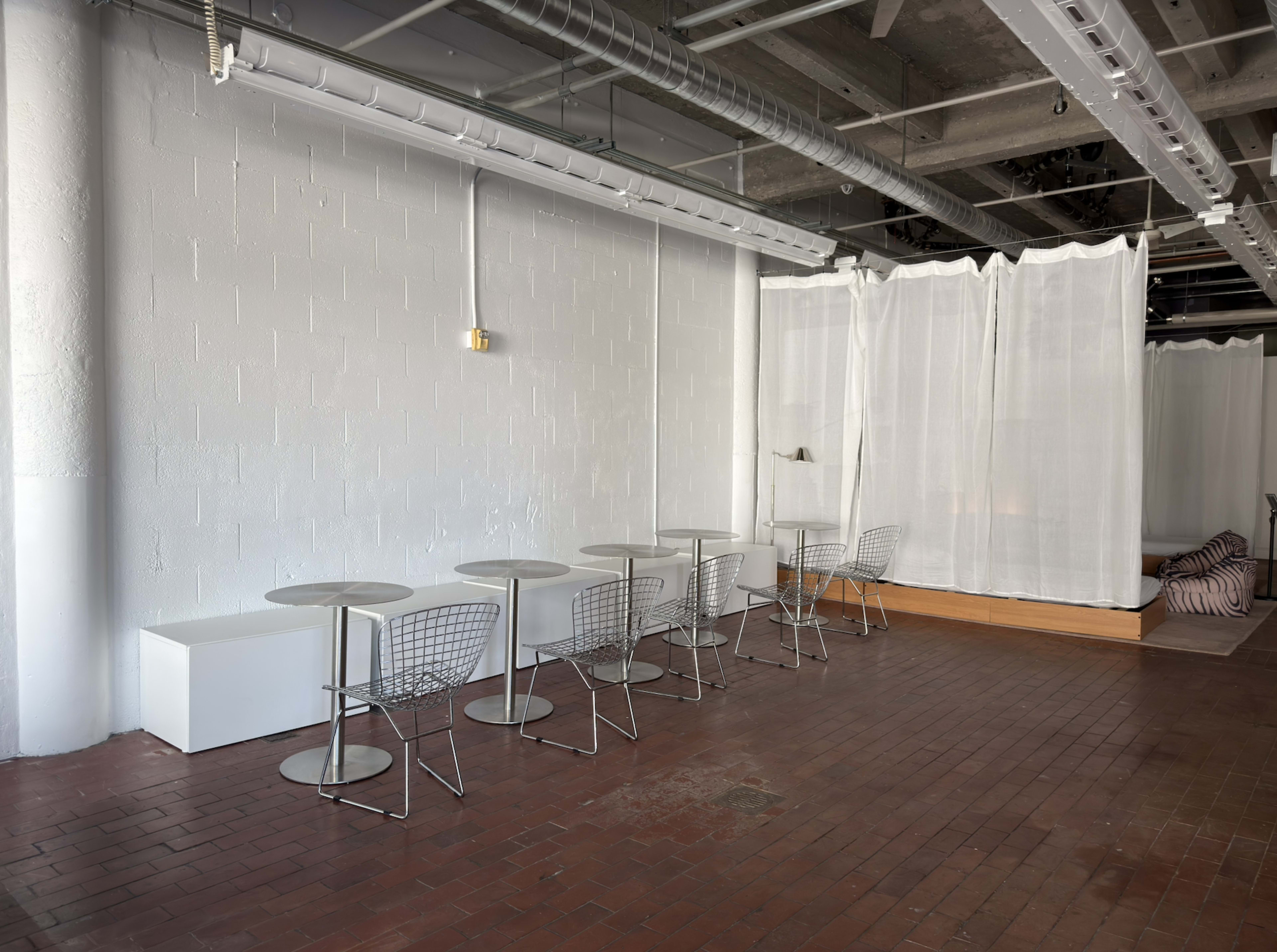 The image shows an empty room with several small metal-topped tables and wire chairs arranged against a white wall, with a partition of sheer curtains at the back.