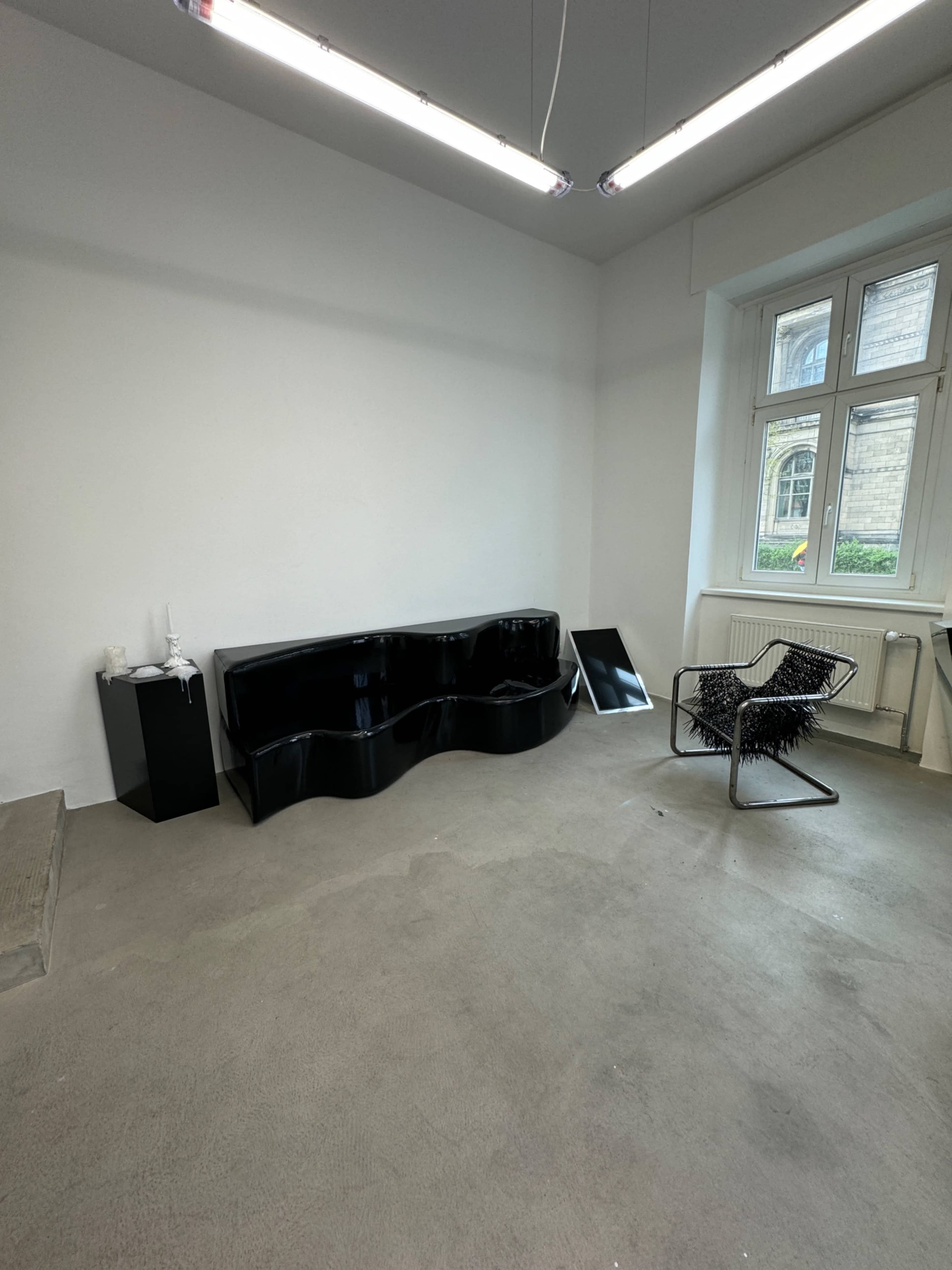 The image shows a sparsely furnished room with a black wave-shaped couch, a black square table, a reflective panel, and a chair with a wire frame.