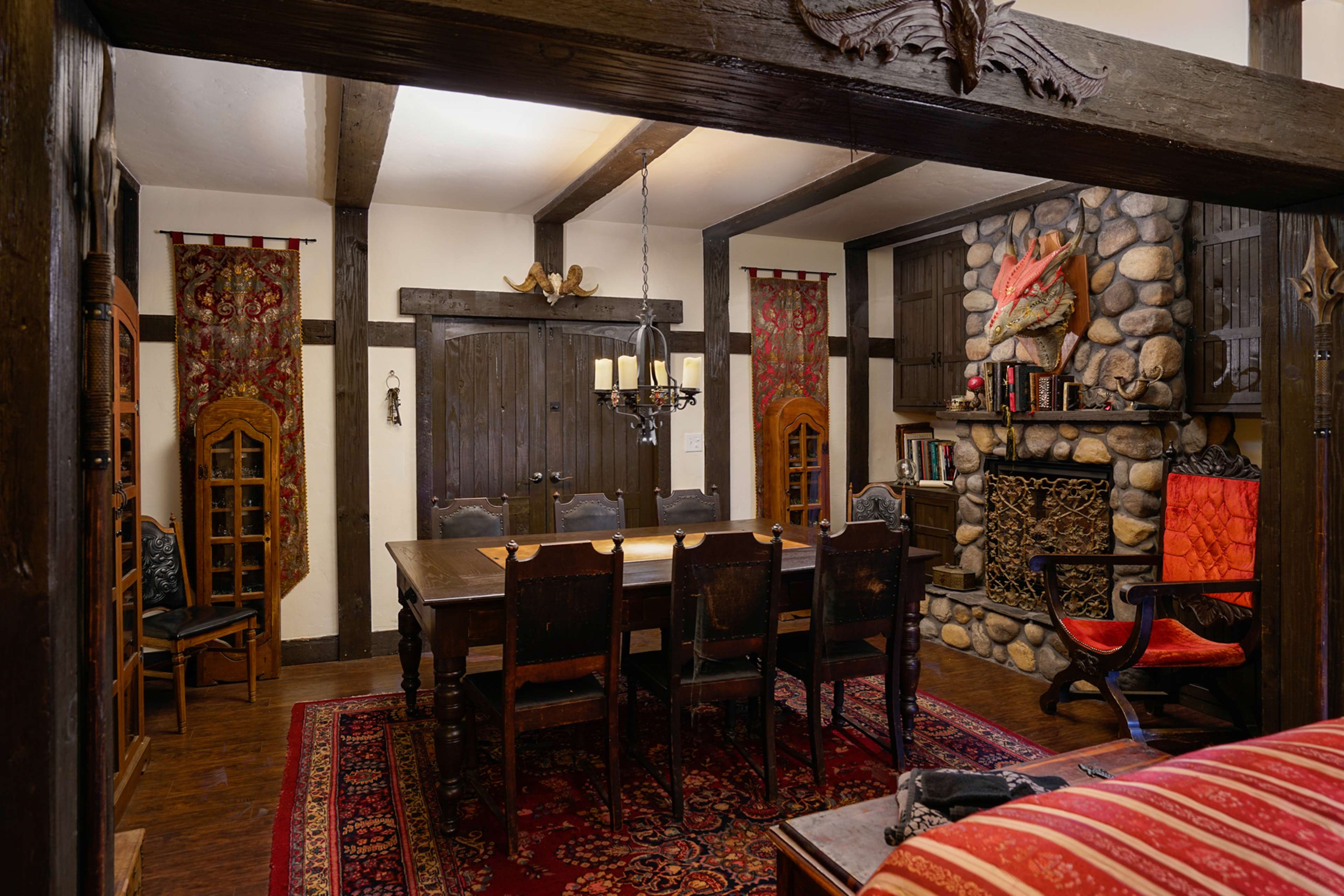 The image shows a rustic dining room featuring a large wooden table surrounded by chairs, a stone fireplace, and decorative wall hangings.