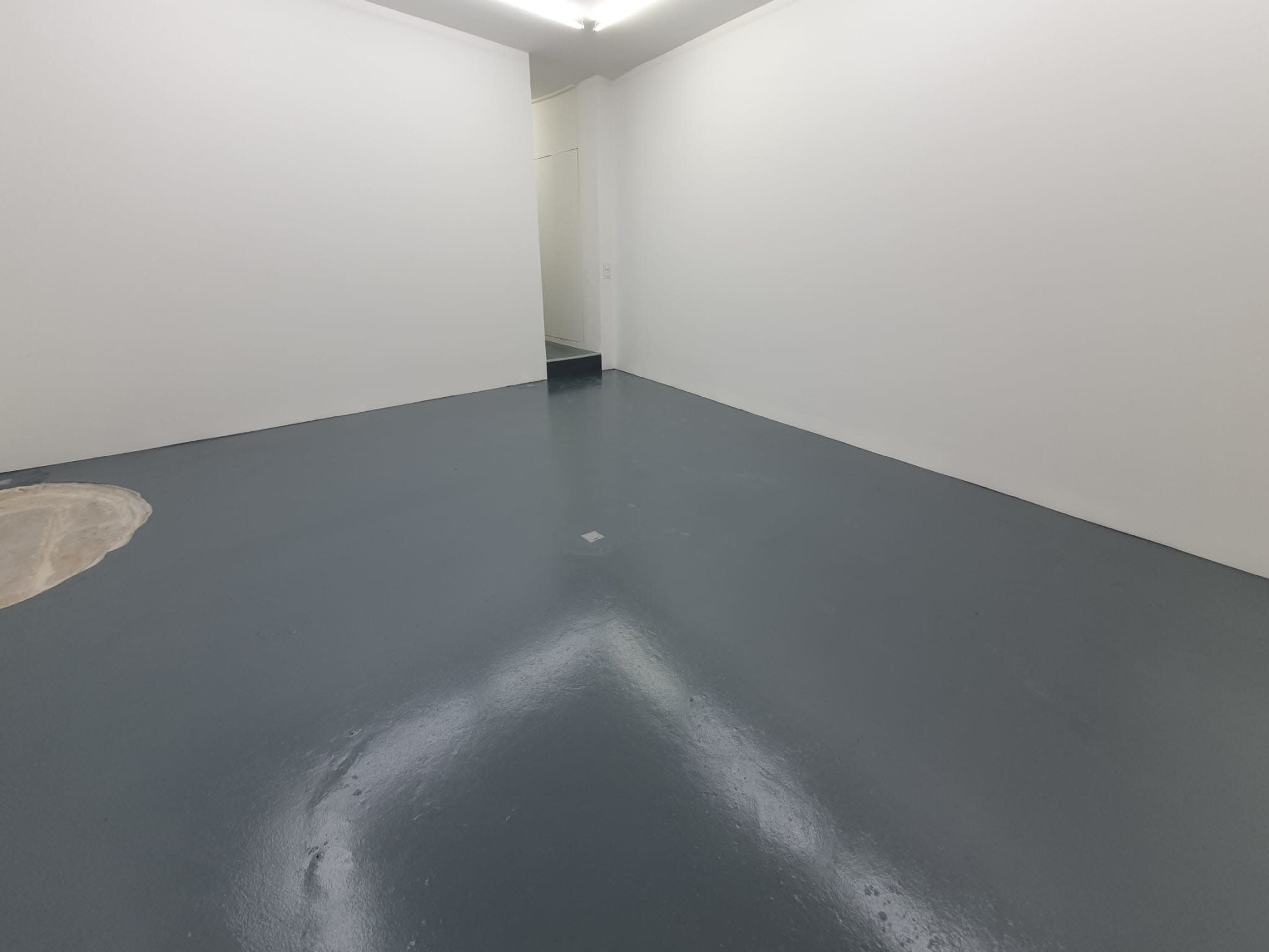 A room with a freshly painted gray floor and plain white walls, featuring a small, circular patch of unfinished flooring in the corner.