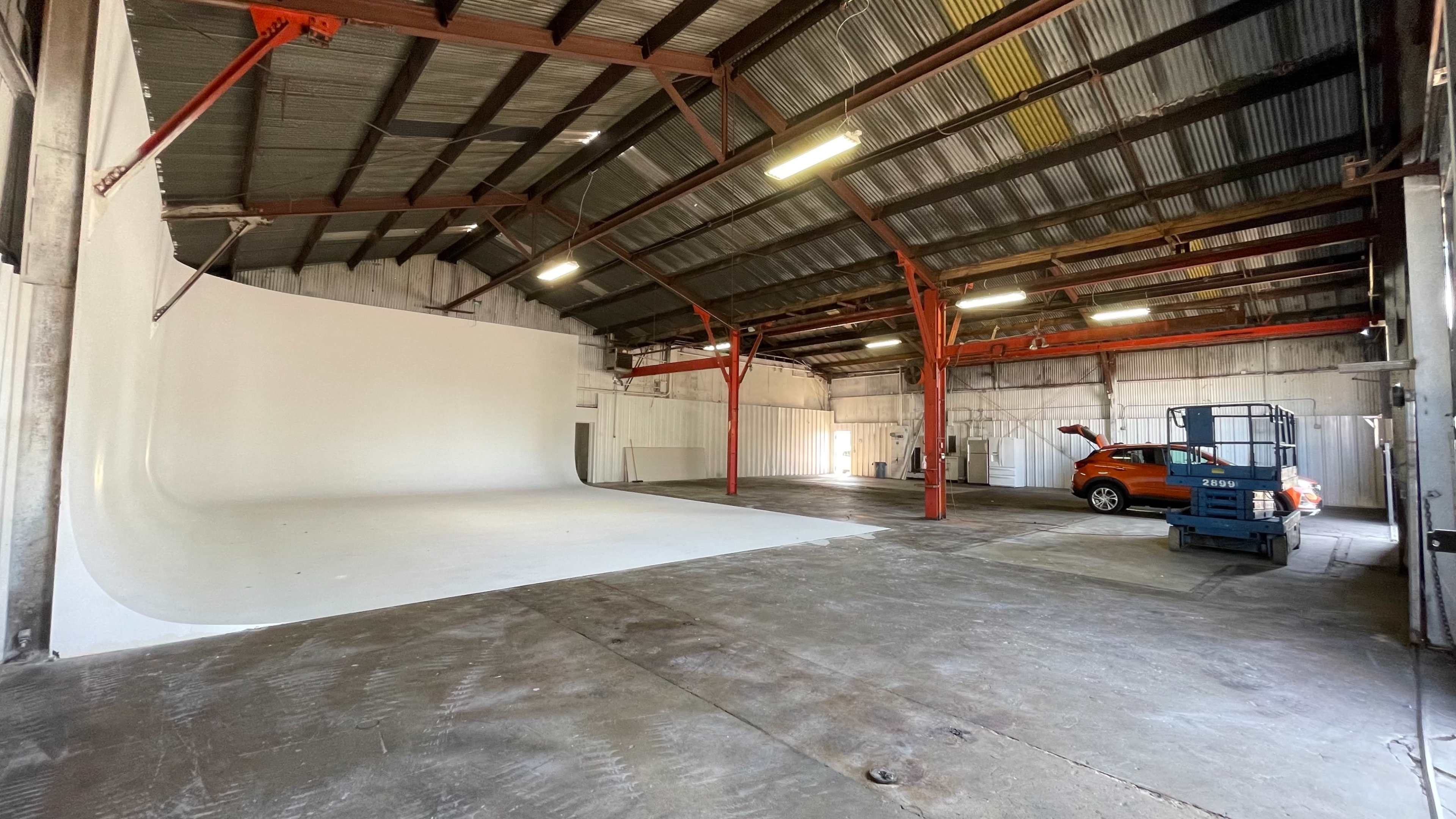 An industrial warehouse space with a large white backdrop and a cherry picker alongside a parked orange vehicle.