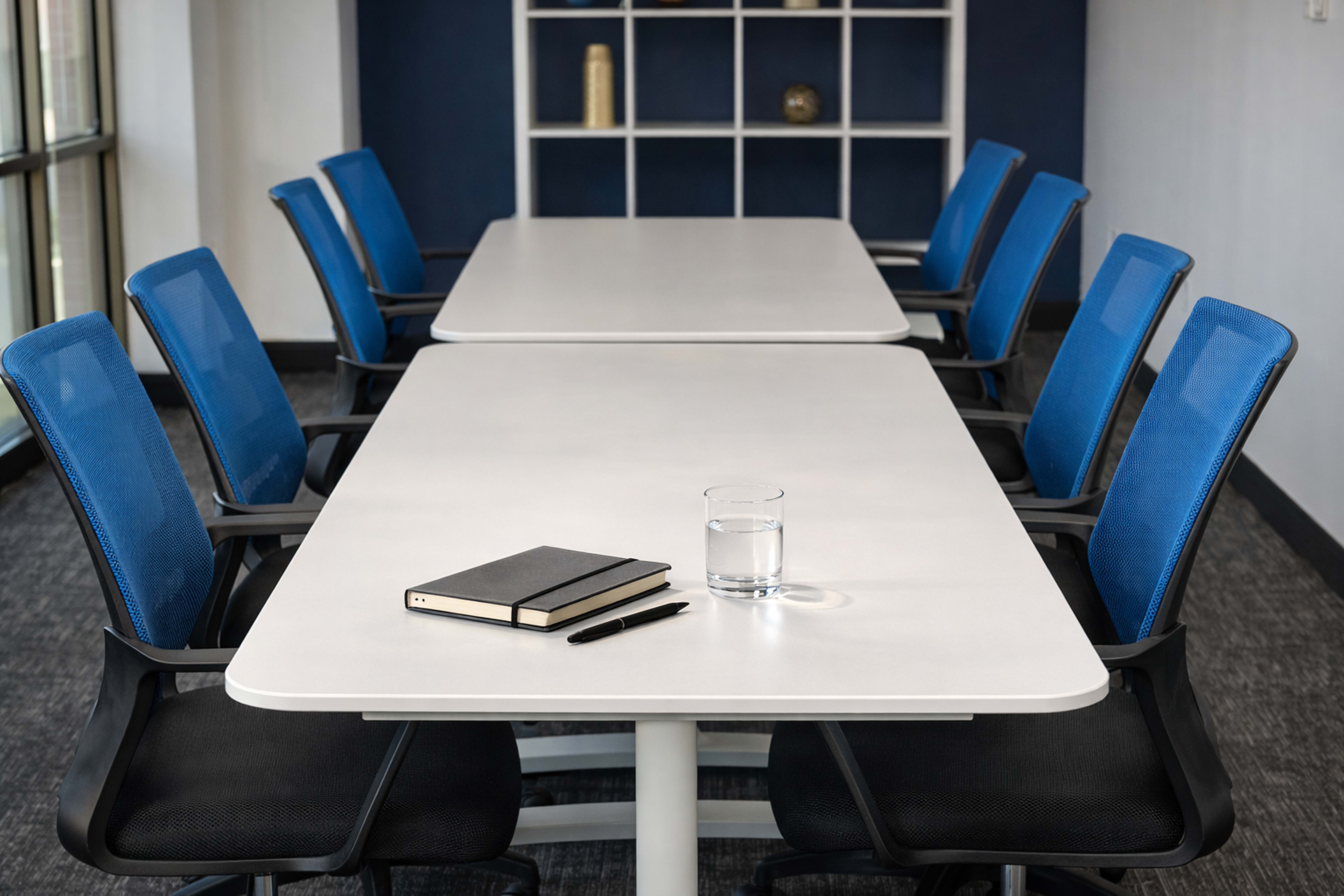 A modern conference room features two long rectangular tables facing each other, with blue mesh chairs and a glass of water, a notebook, and a pen on one of the tables.