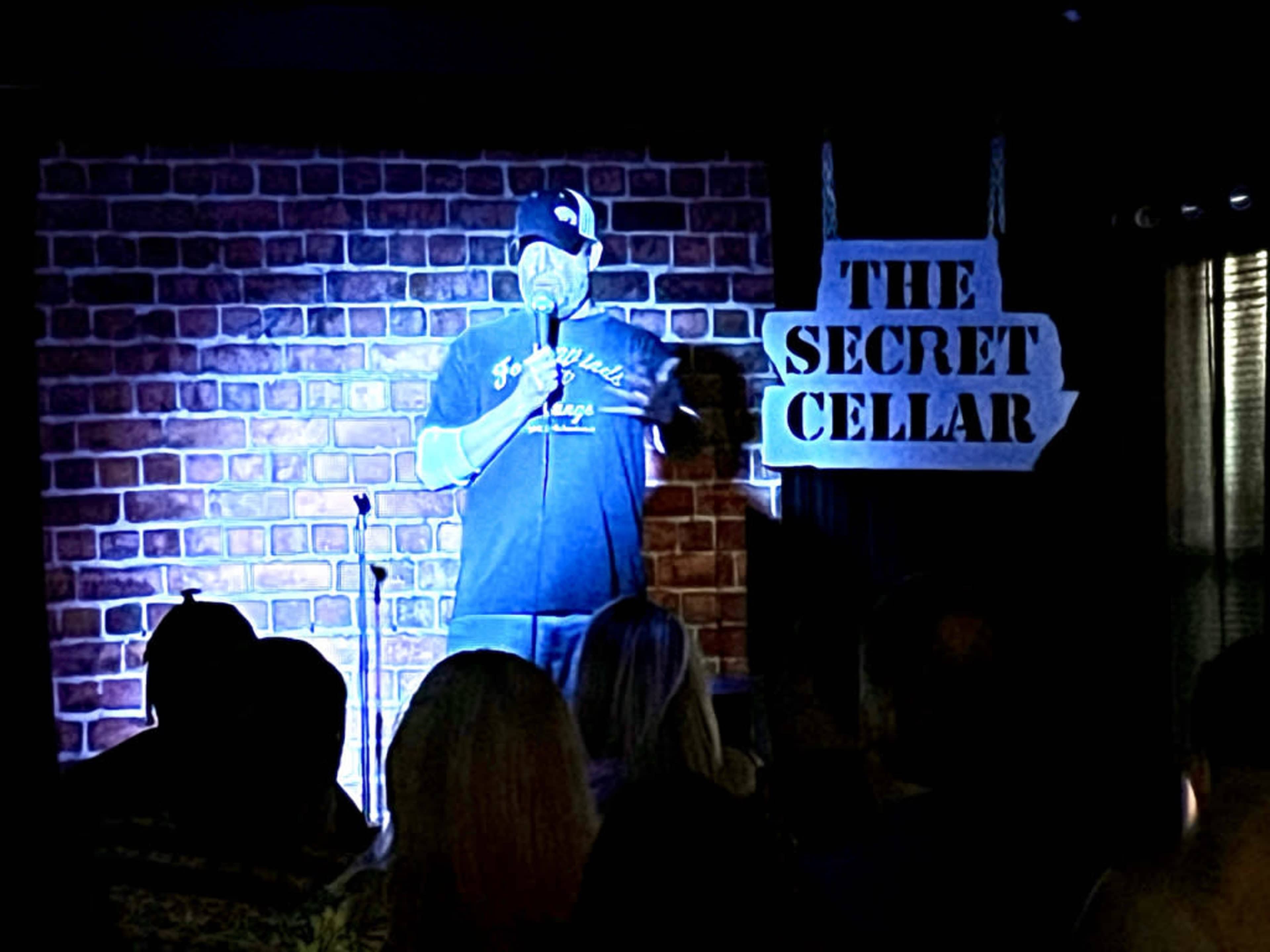 A comedian performs on stage in front of an audience at a venue called "The Secret Cellar," with a brick wall backdrop.