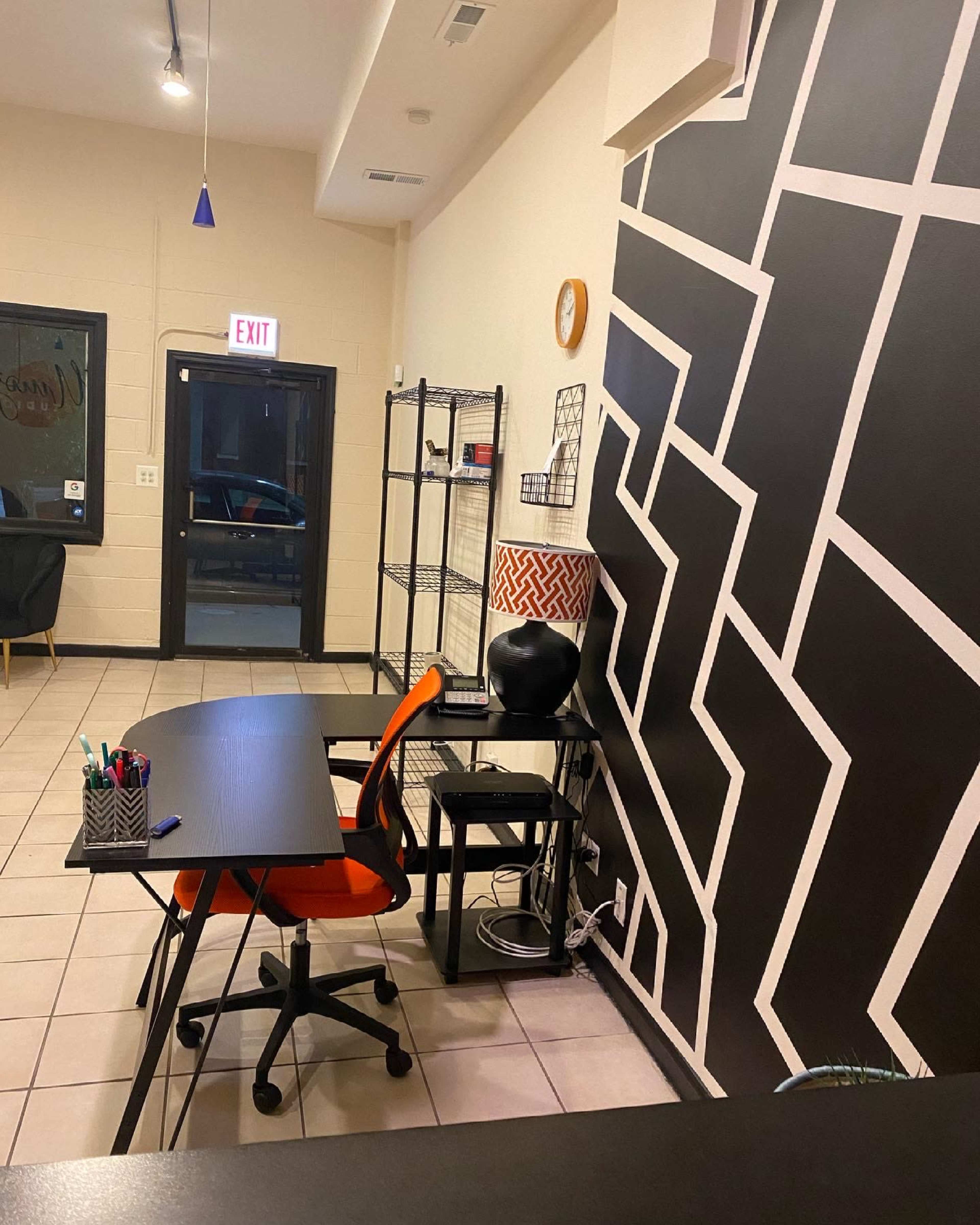 An interior workspace with a black and white geometric patterned wall, a desk with an orange chair, and shelving in the corner.