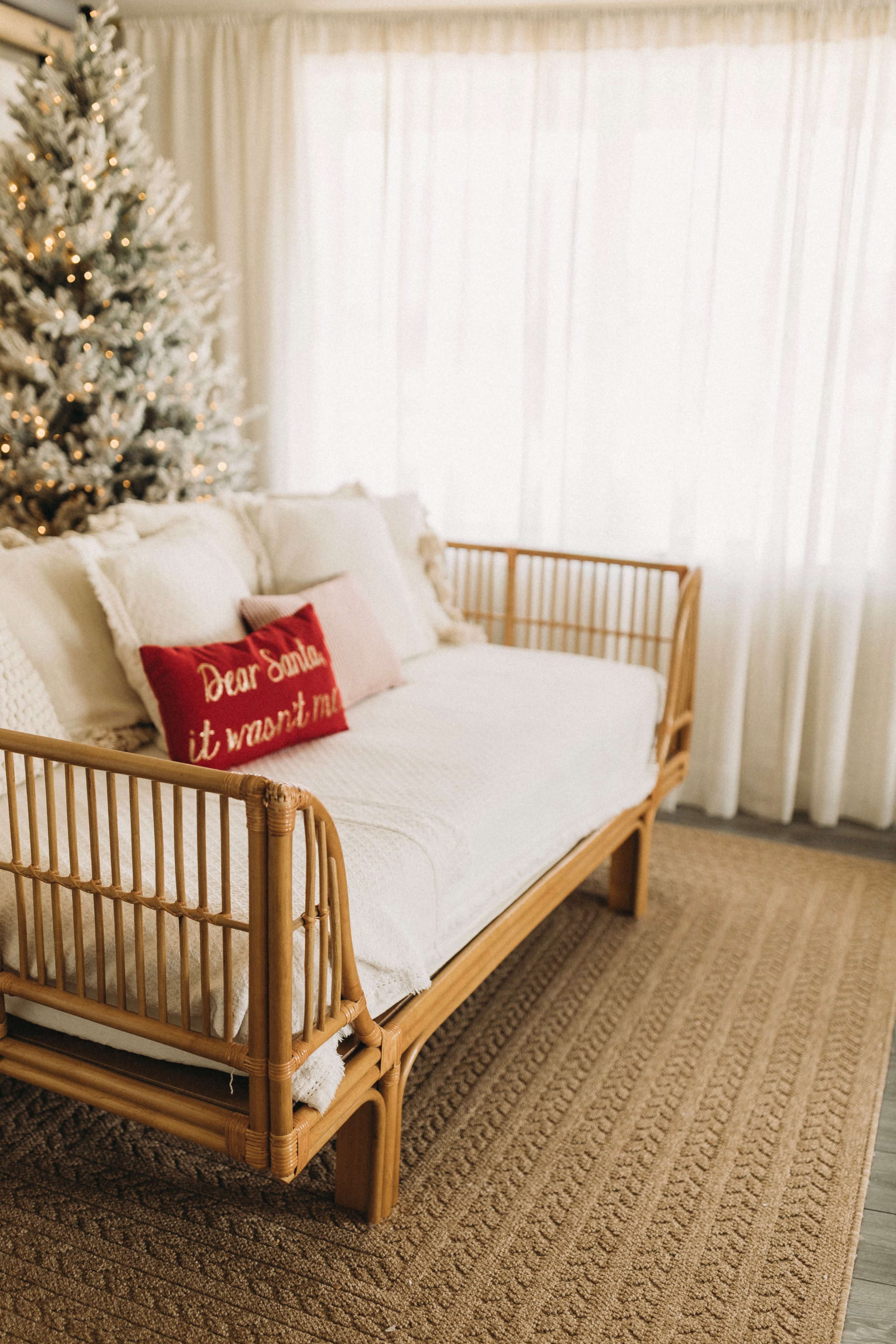 A rattan sofa with white cushions is positioned in front of a tall Christmas tree, with sheer curtains allowing natural light to filter into the room.