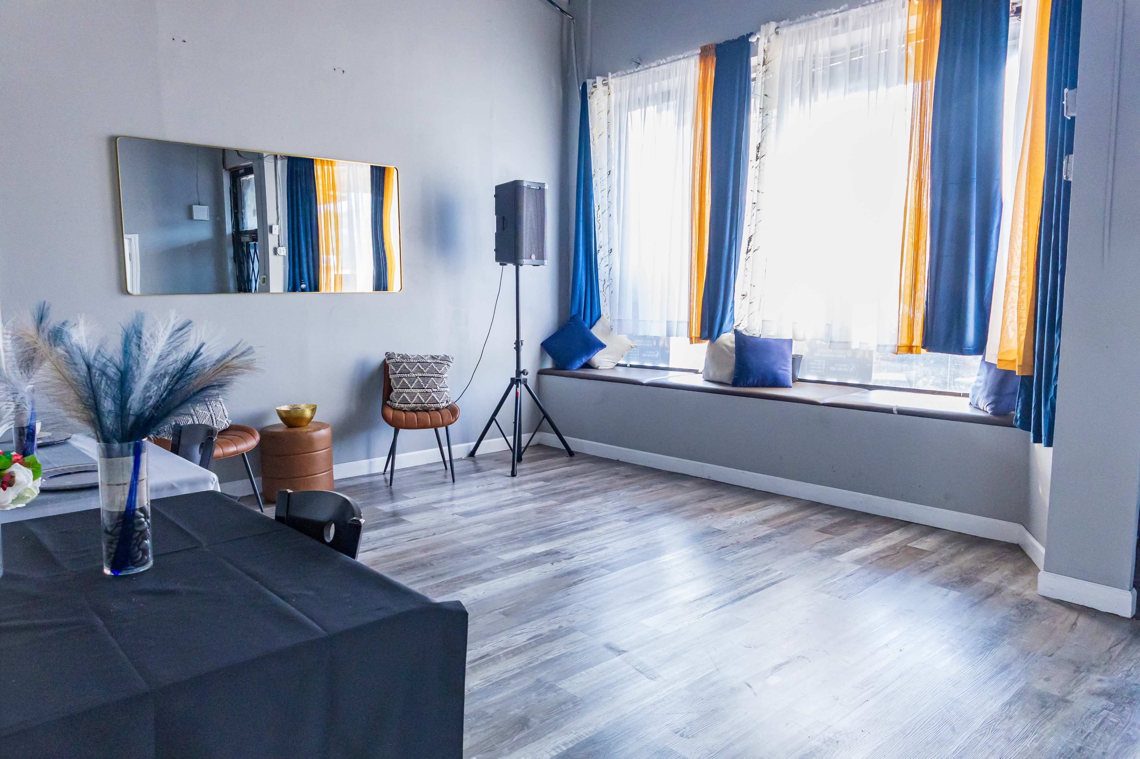 The room features a large mirror, a seating area with cushions by the window, and a small table set against a wall with gray and blue decor.