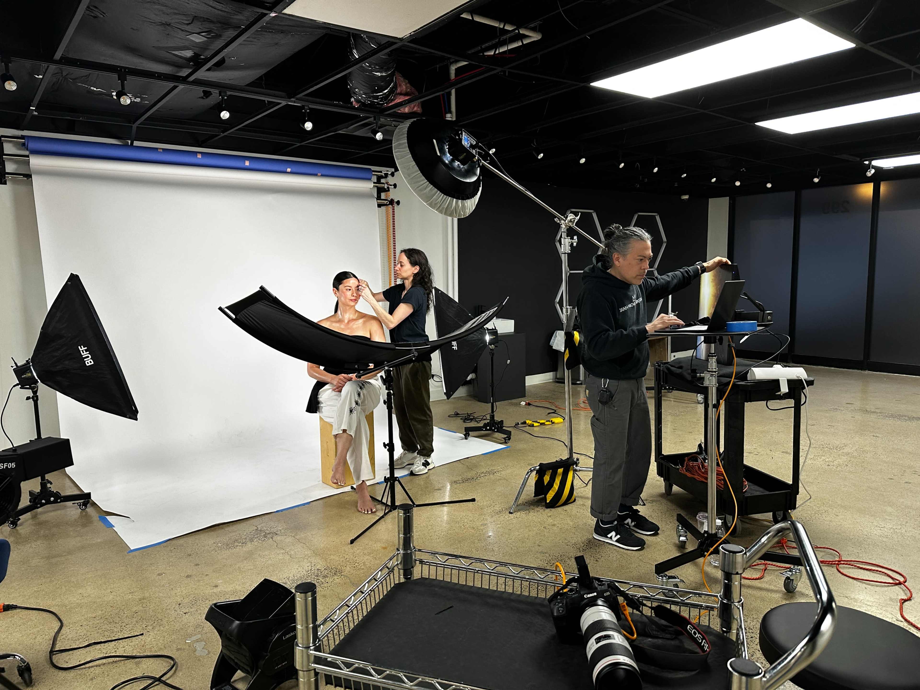 A model poses in front of a white backdrop while a photographer and assistant work on lighting and equipment in a studio setting.