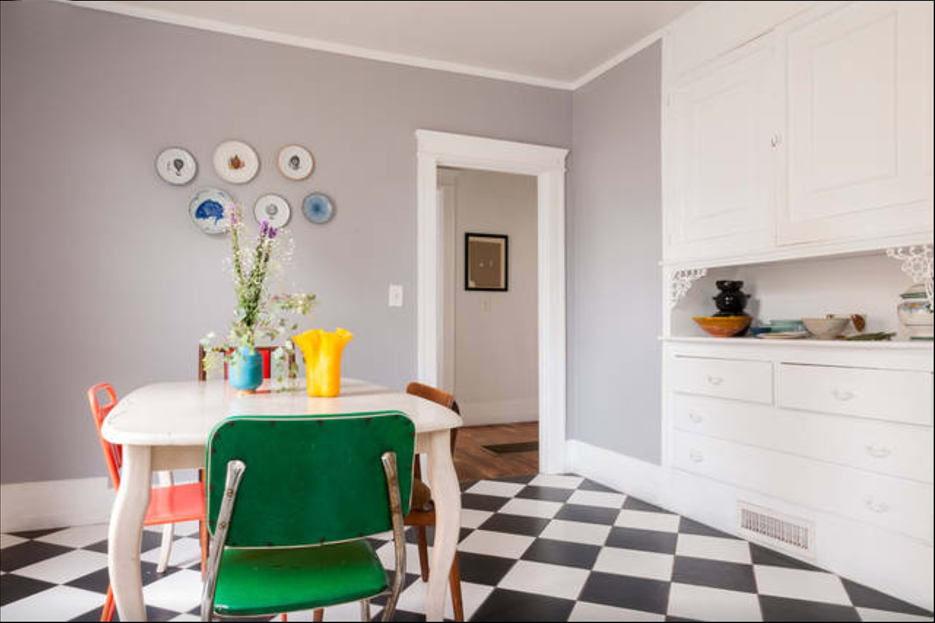 A dining area features a table surrounded by colorful chairs, with decorative plates on the gray wall and a side cabinet displaying dishes.