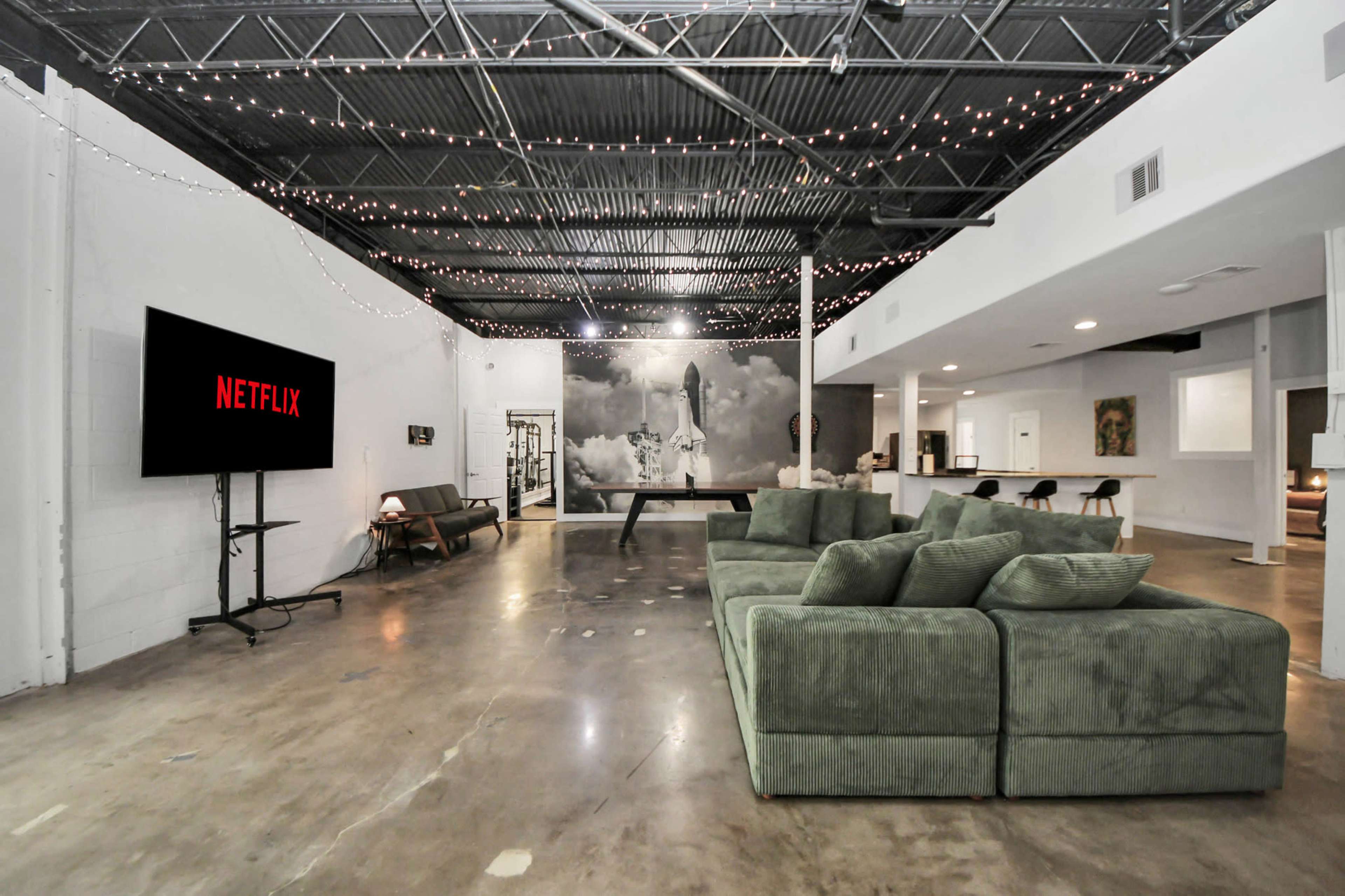 The image shows a spacious, industrial-style living area with a large green sectional couch, a wall-mounted TV displaying the Netflix logo, and casual dining and seating arrangements in a well-lit environment.