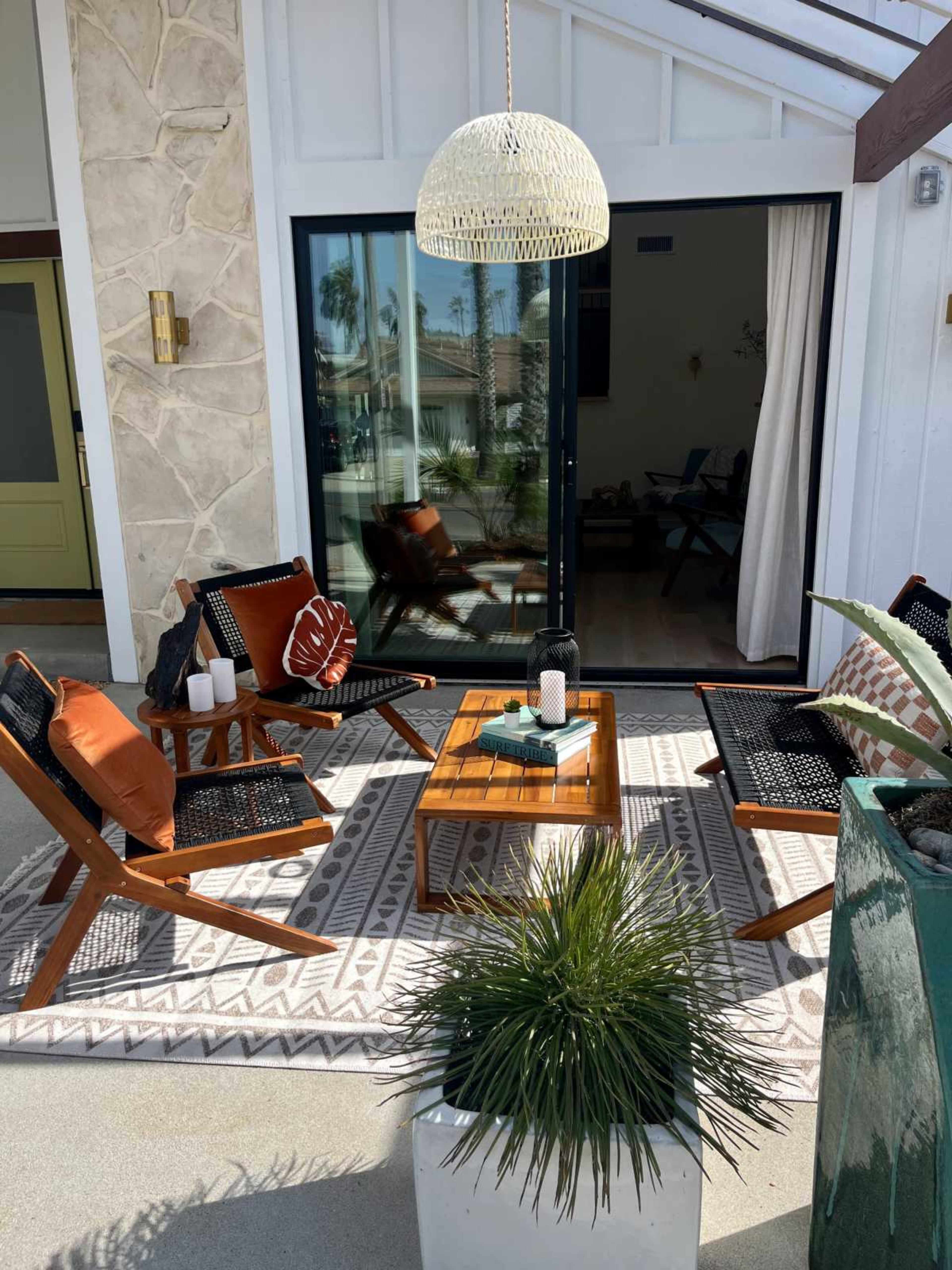 A modern outdoor seating area features wicker chairs with cushions, a wooden coffee table, and a large pendant light, surrounded by potted plants and a patterned area rug.