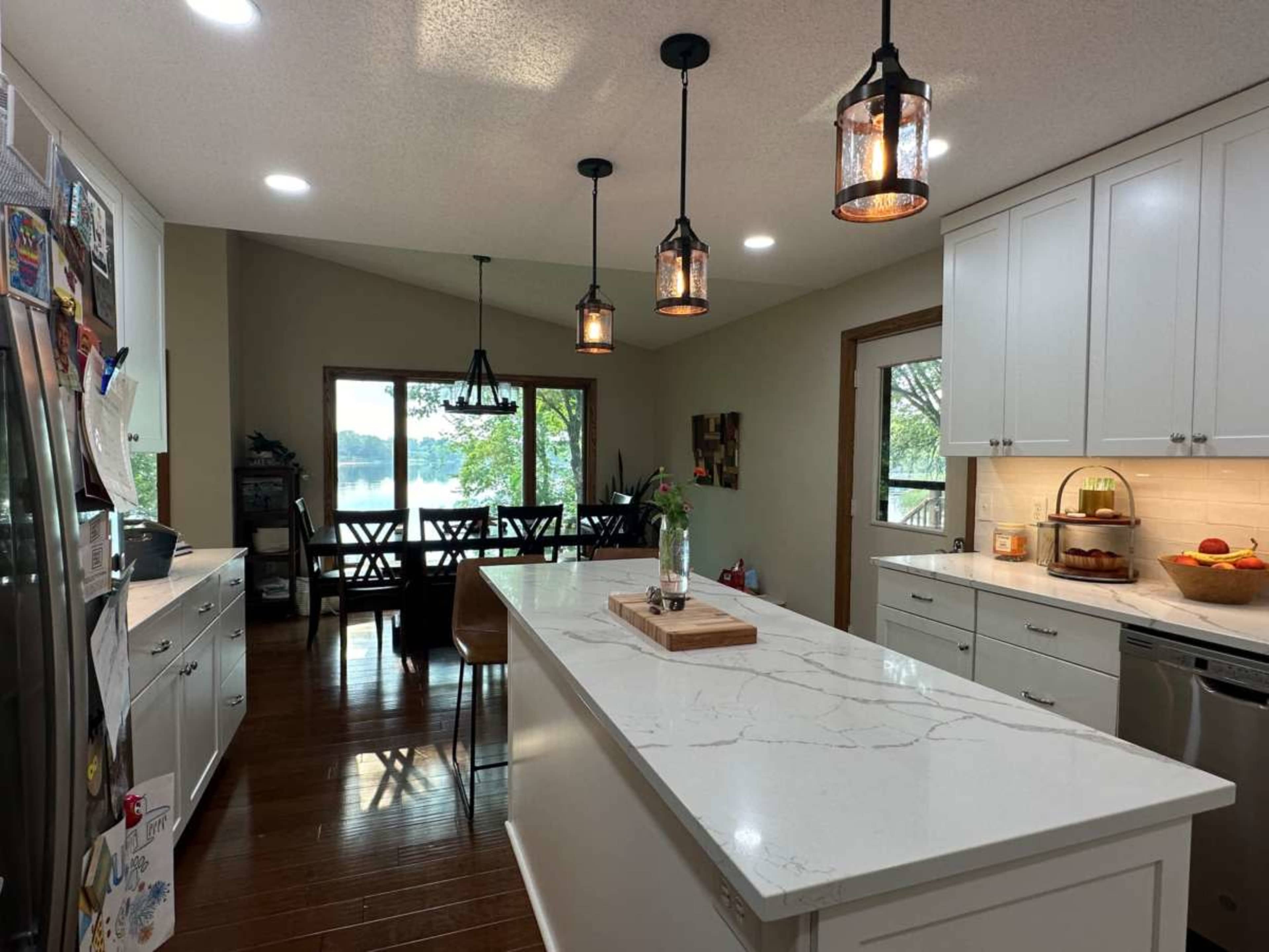 The image shows a modern kitchen with white cabinetry, a large marble island, and pendant lighting, overlooking a dining area with a view of a lake through large windows.