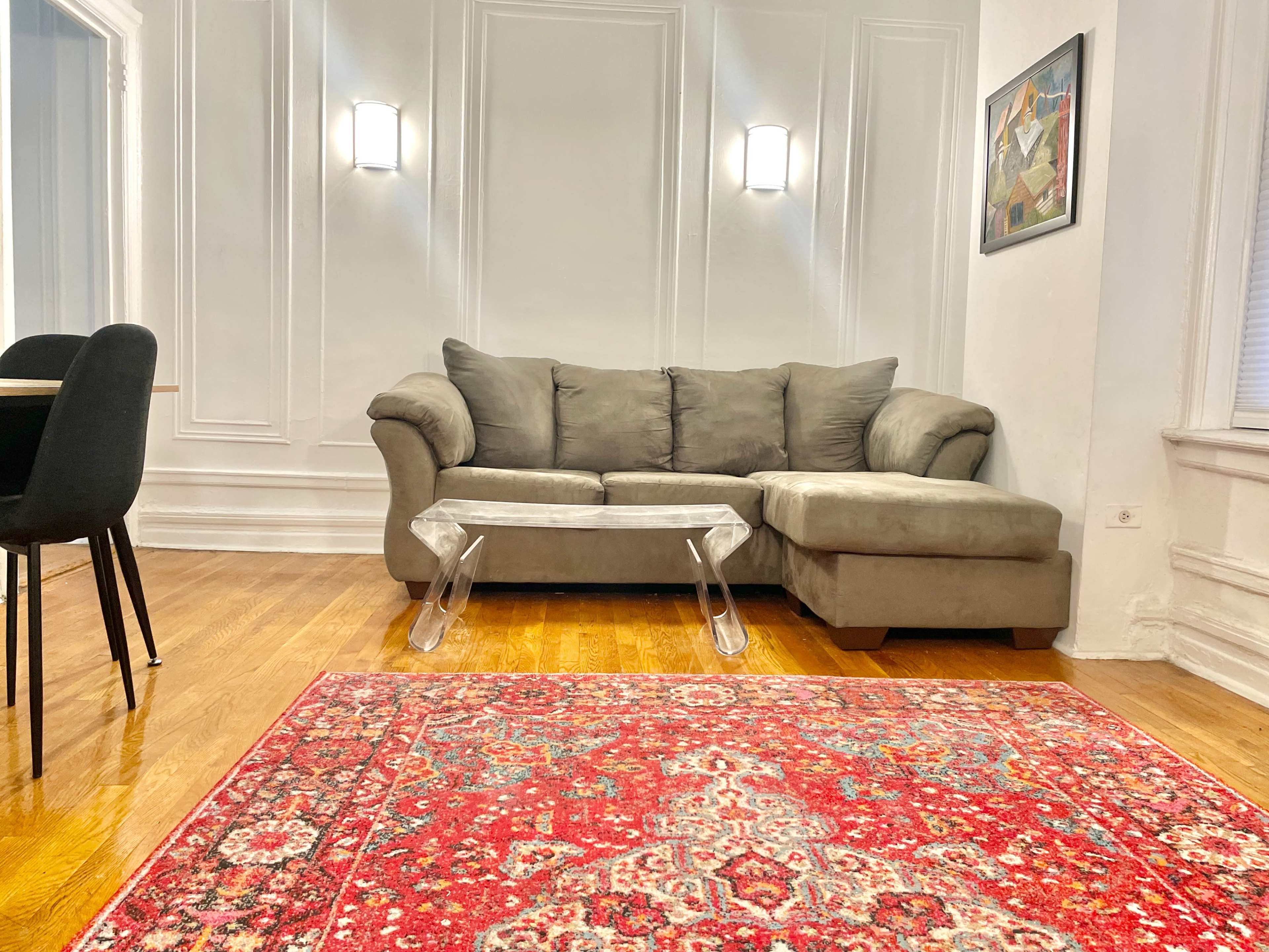 A living room with a gray sectional sofa, a clear acrylic coffee table, and a red patterned rug on wooden flooring.