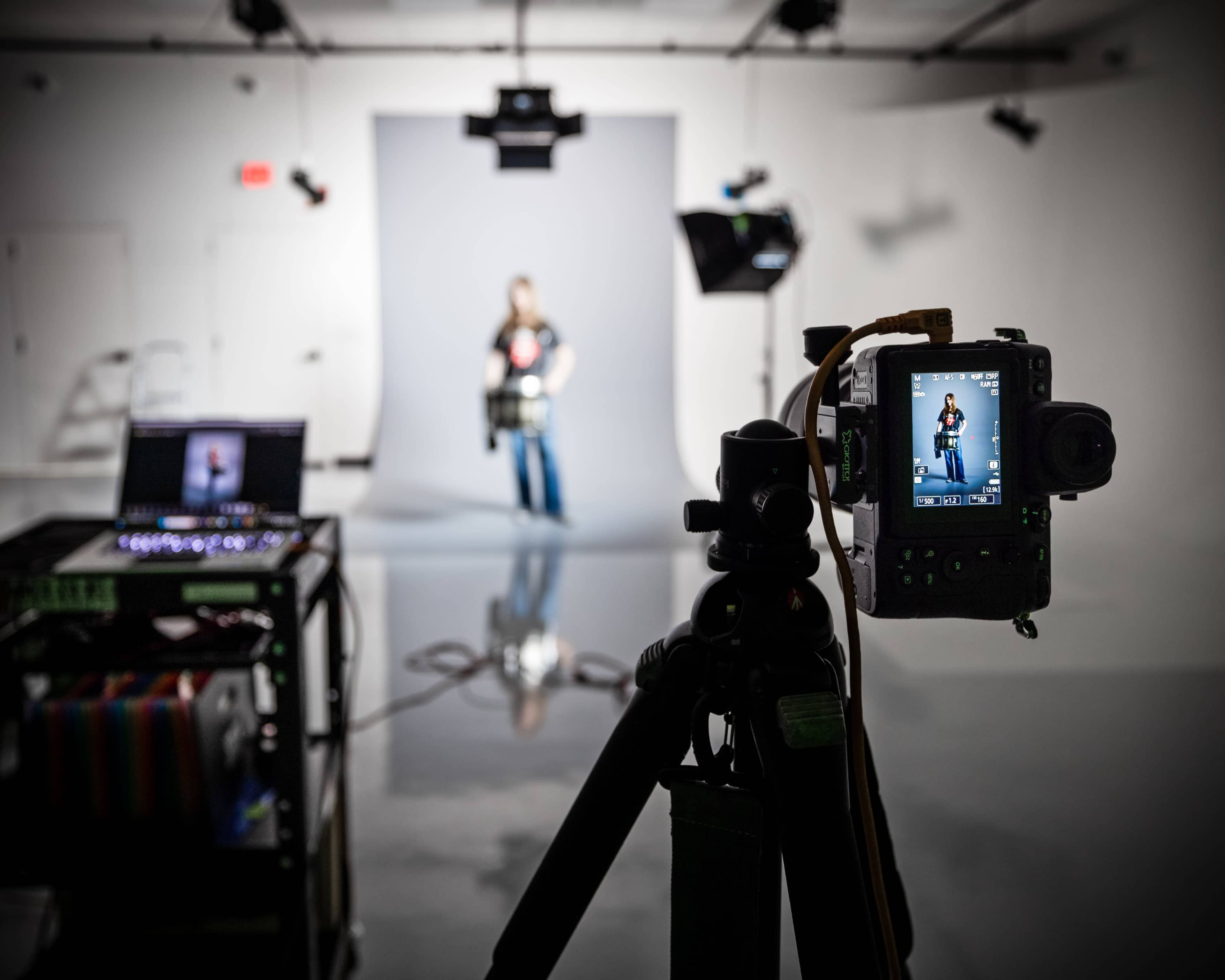 A camera on a tripod captures a model posing in front of a gray backdrop in a studio setting.