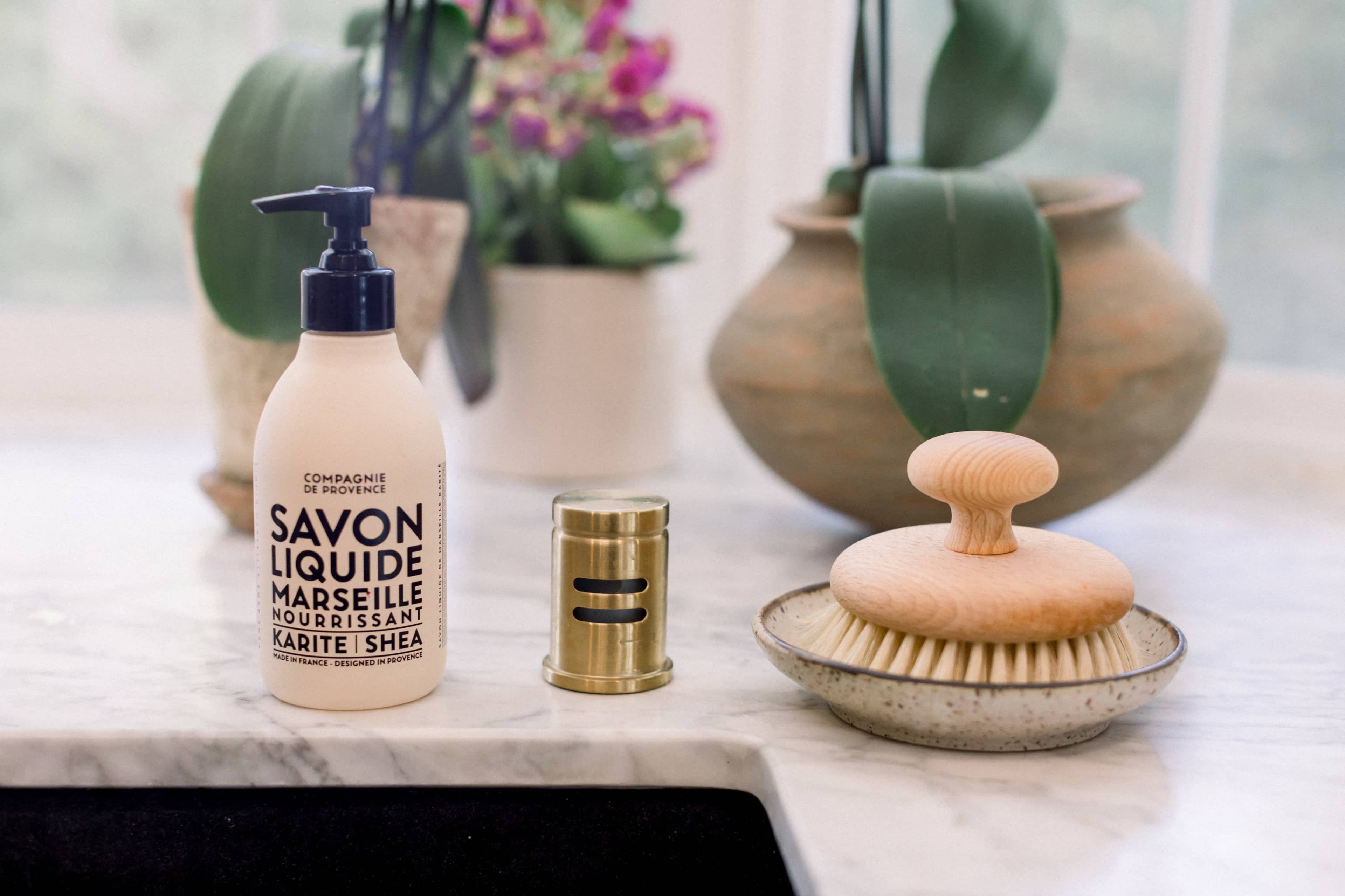A bottle of liquid soap, a wooden brush, and a metal salt shaker are arranged on a marble countertop, with plants visible in the background.