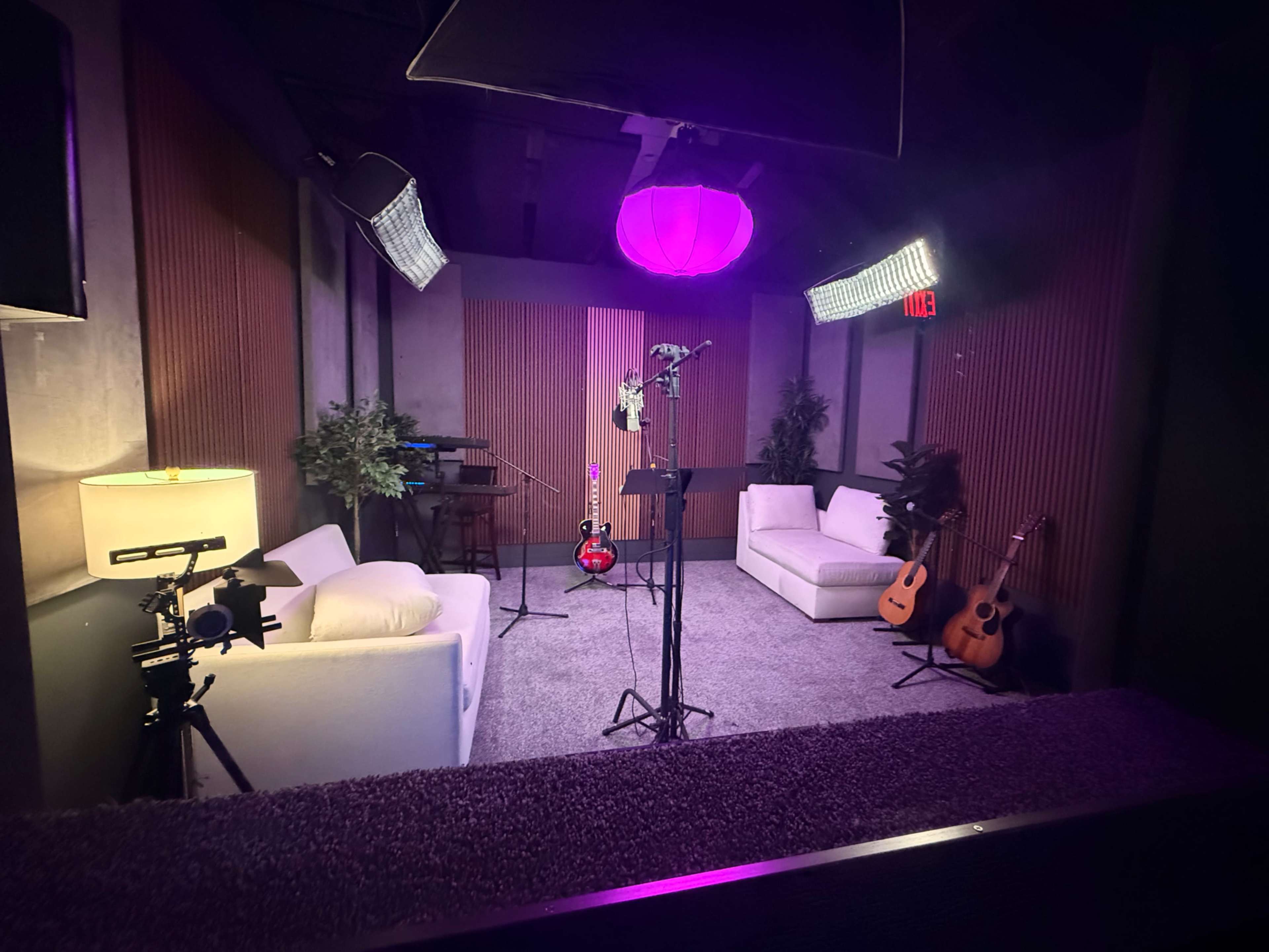 The image shows a studio setup with two white couches, a microphone stand, guitars, and soft lighting.