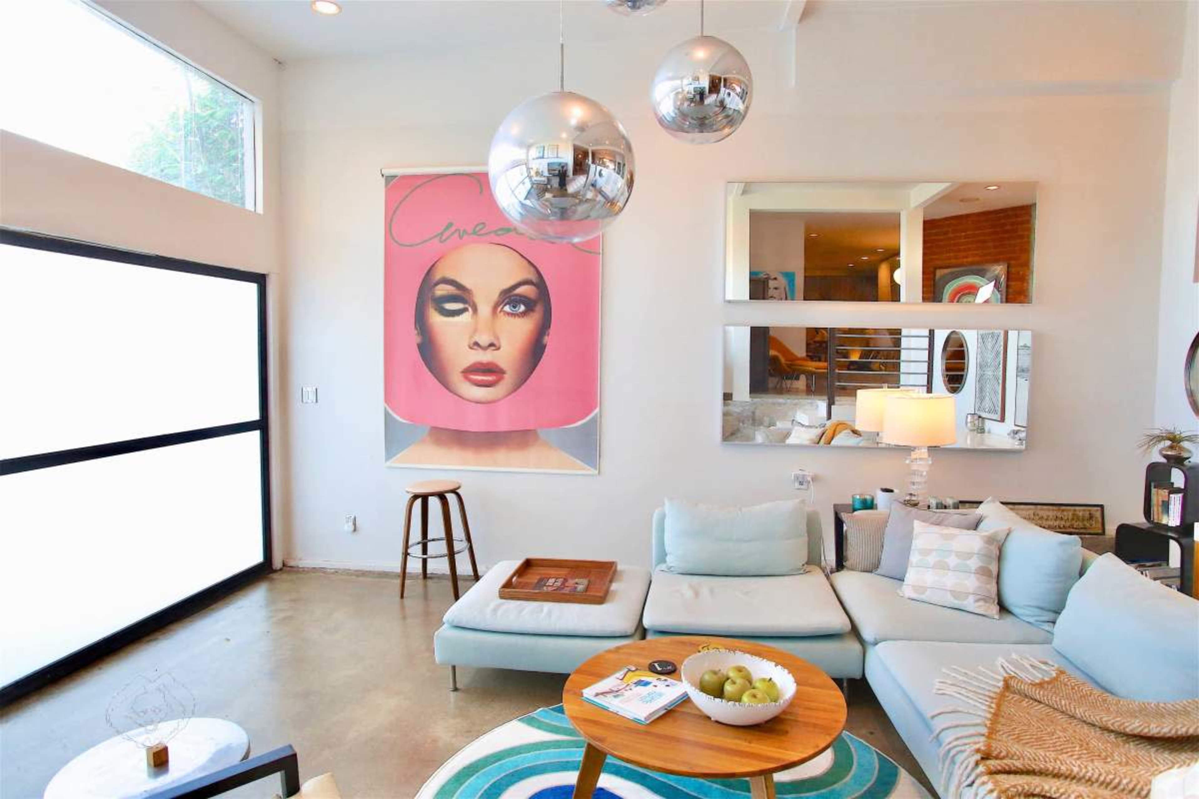 The image shows a modern living room featuring a light blue sectional sofa, a round wooden coffee table with decorative items, and a large art piece hanging on the wall.