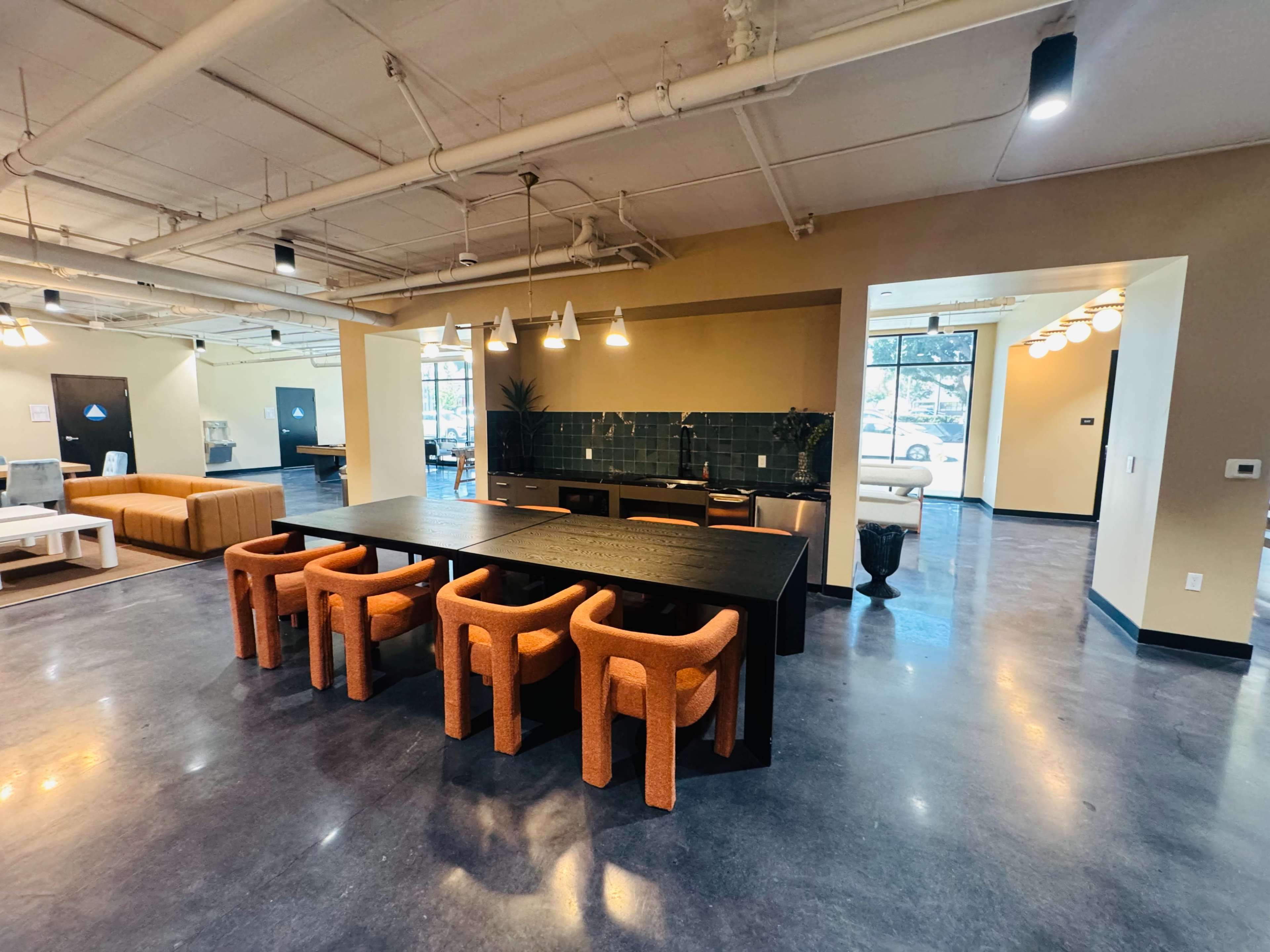 The image shows a spacious indoor area with a long table surrounded by orange chairs, a kitchenette in the background, and various seating arrangements visible throughout the room.