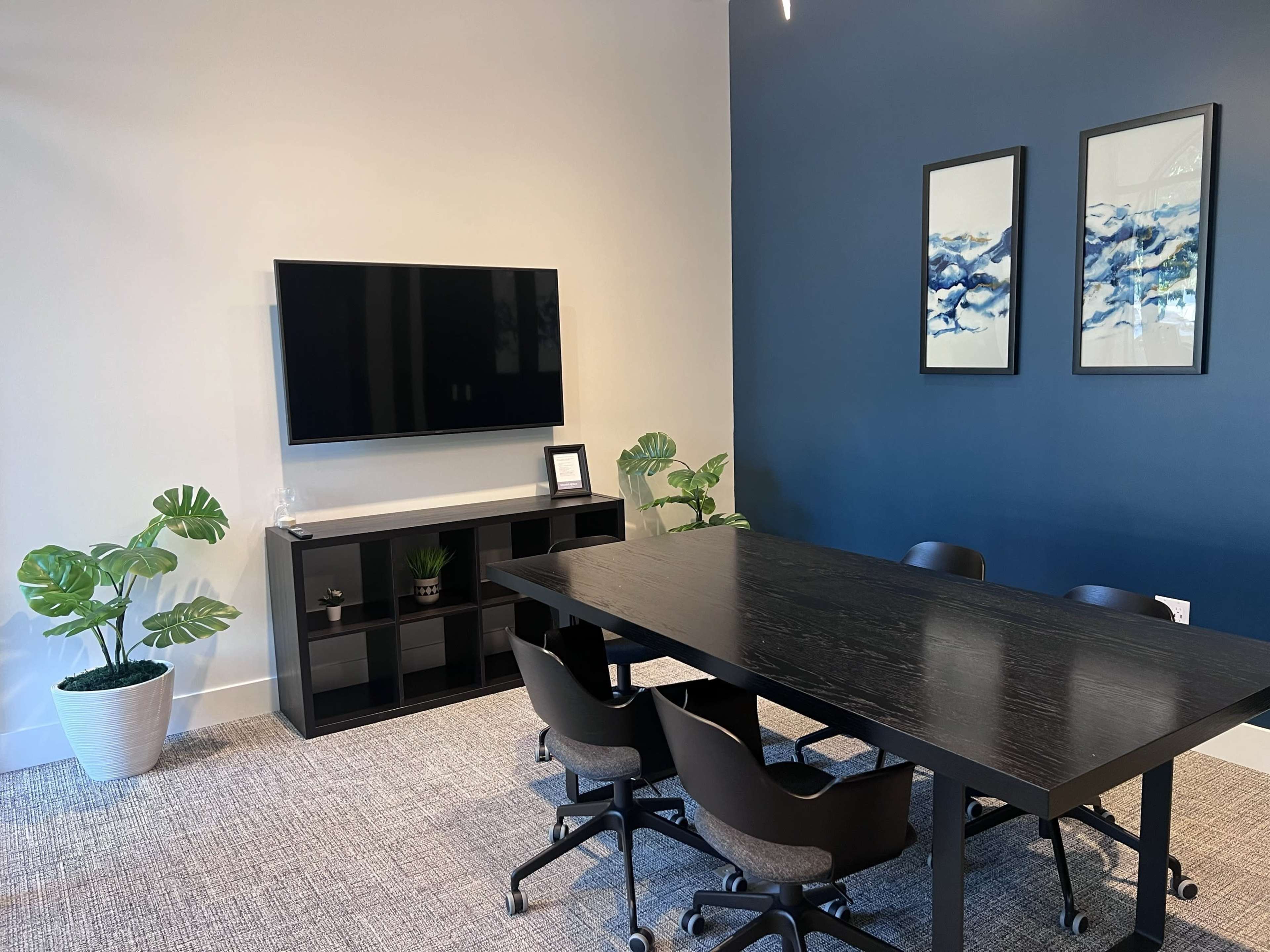 A modern conference room features a large black table, several rolling chairs, a mounted television, and potted plants against a dark blue wall.