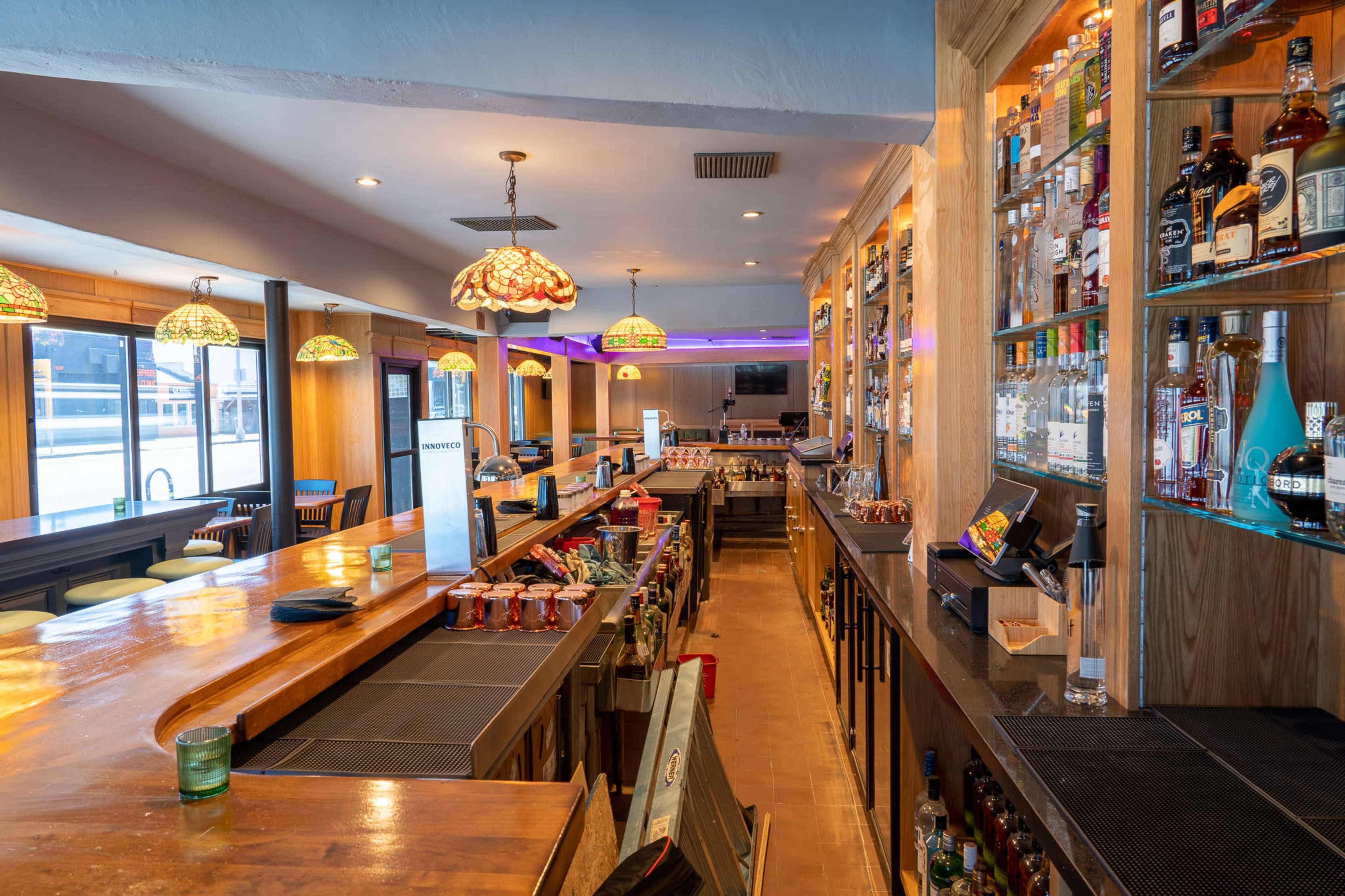A brightly lit bar with a long wooden counter, shelves filled with various bottles of liquor, and colorful pendant lights hanging from the ceiling.