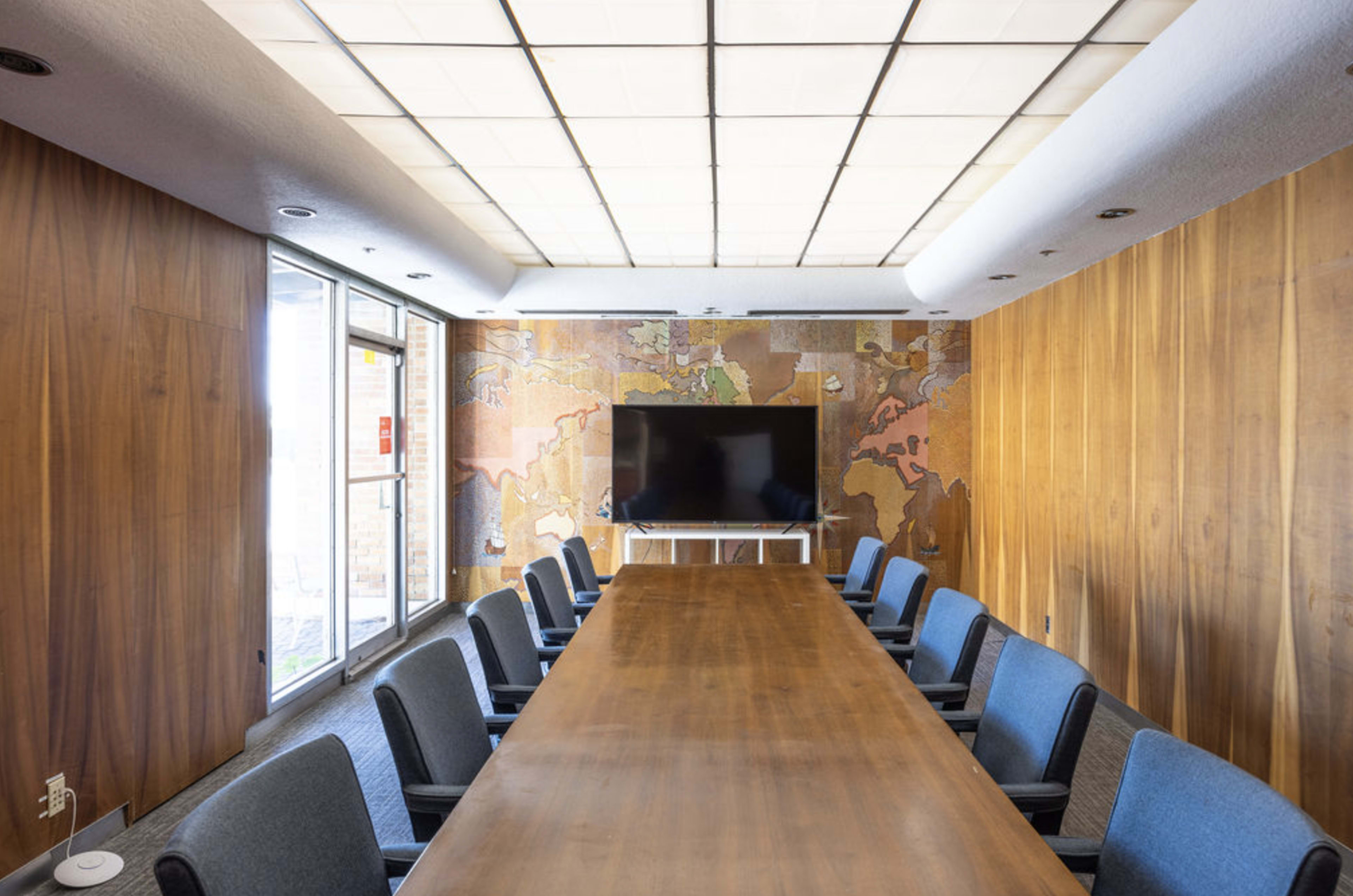 A long wooden conference table is surrounded by gray chairs in a modern meeting room with a large screen and a map mural on the wall.