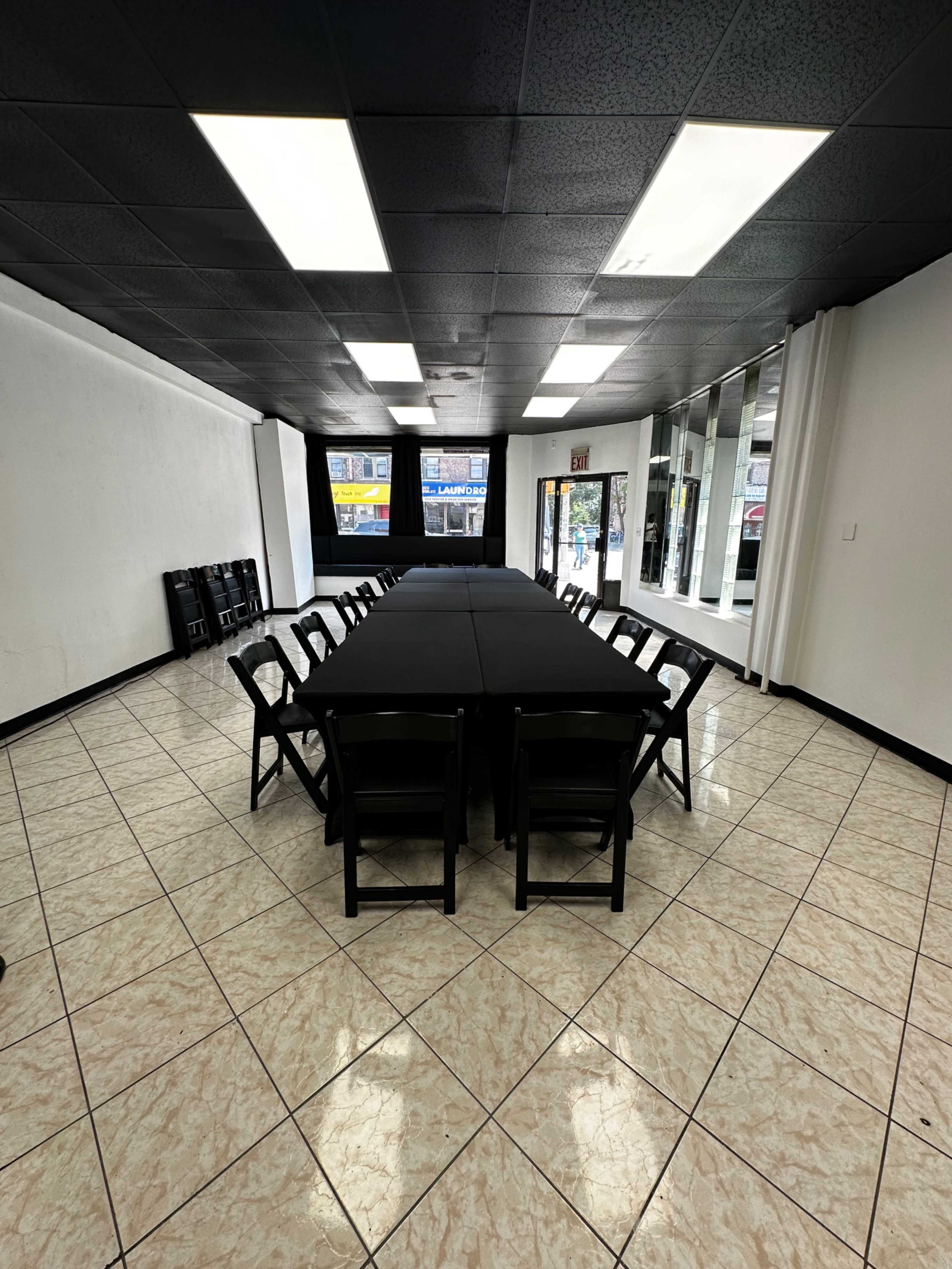 A large, empty room features a long table covered with a black tablecloth surrounded by black chairs, with windows and a doorway along one wall.
