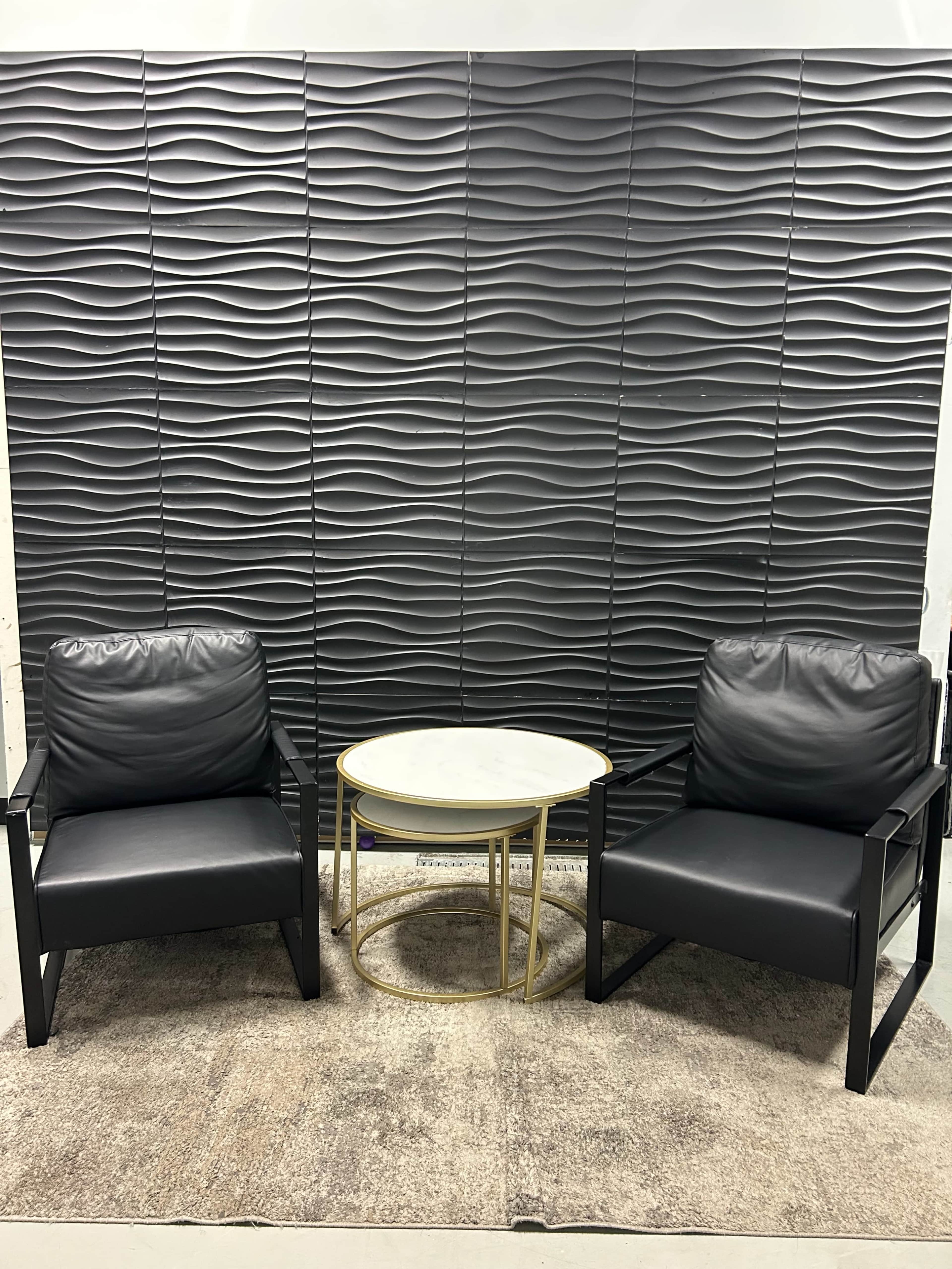 Two black leather chairs face a small round table on a textured gray rug, with a wavy patterned wall in the background.