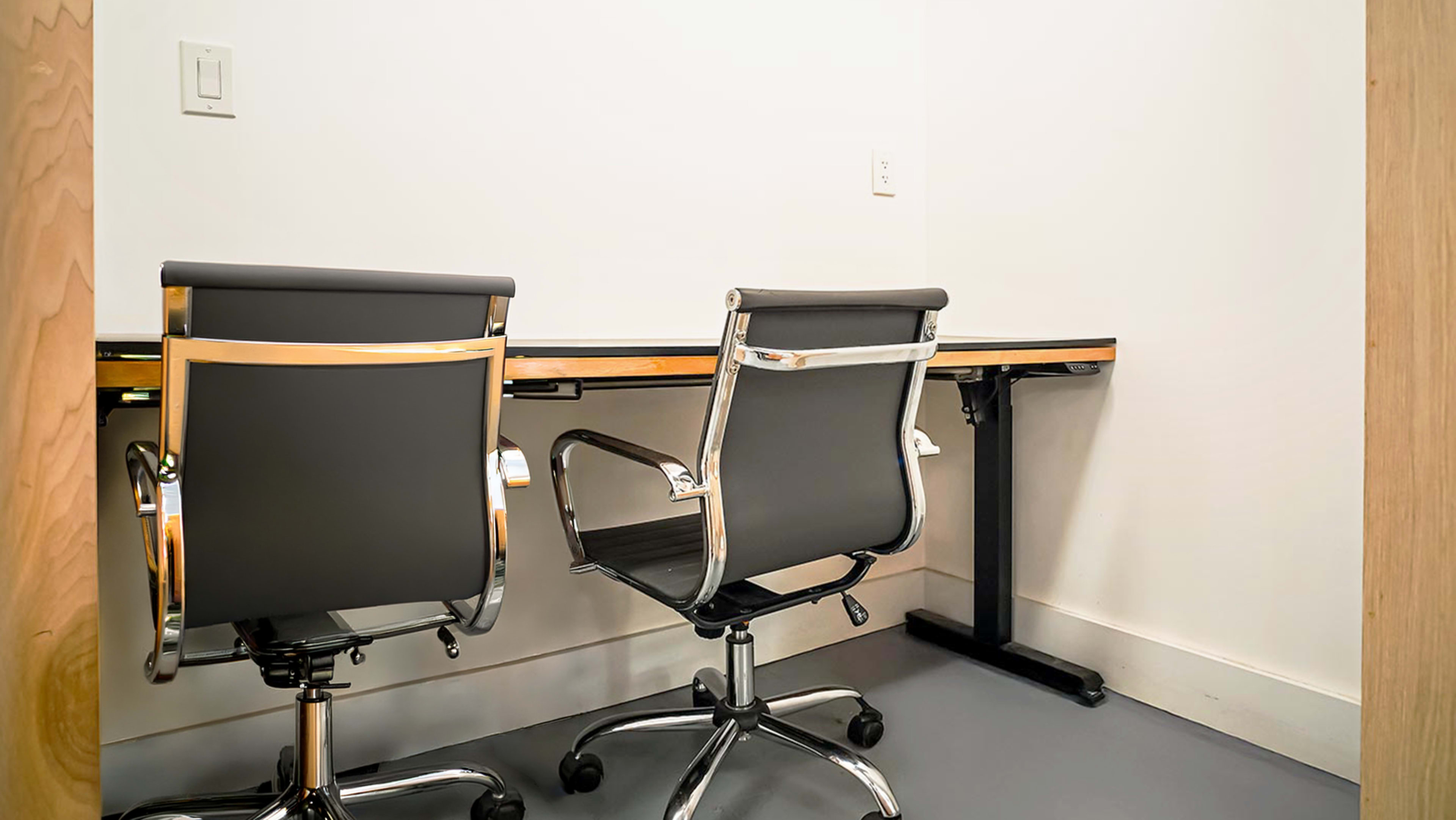 The image shows a small office space with a simple desk and two black office chairs positioned at it.