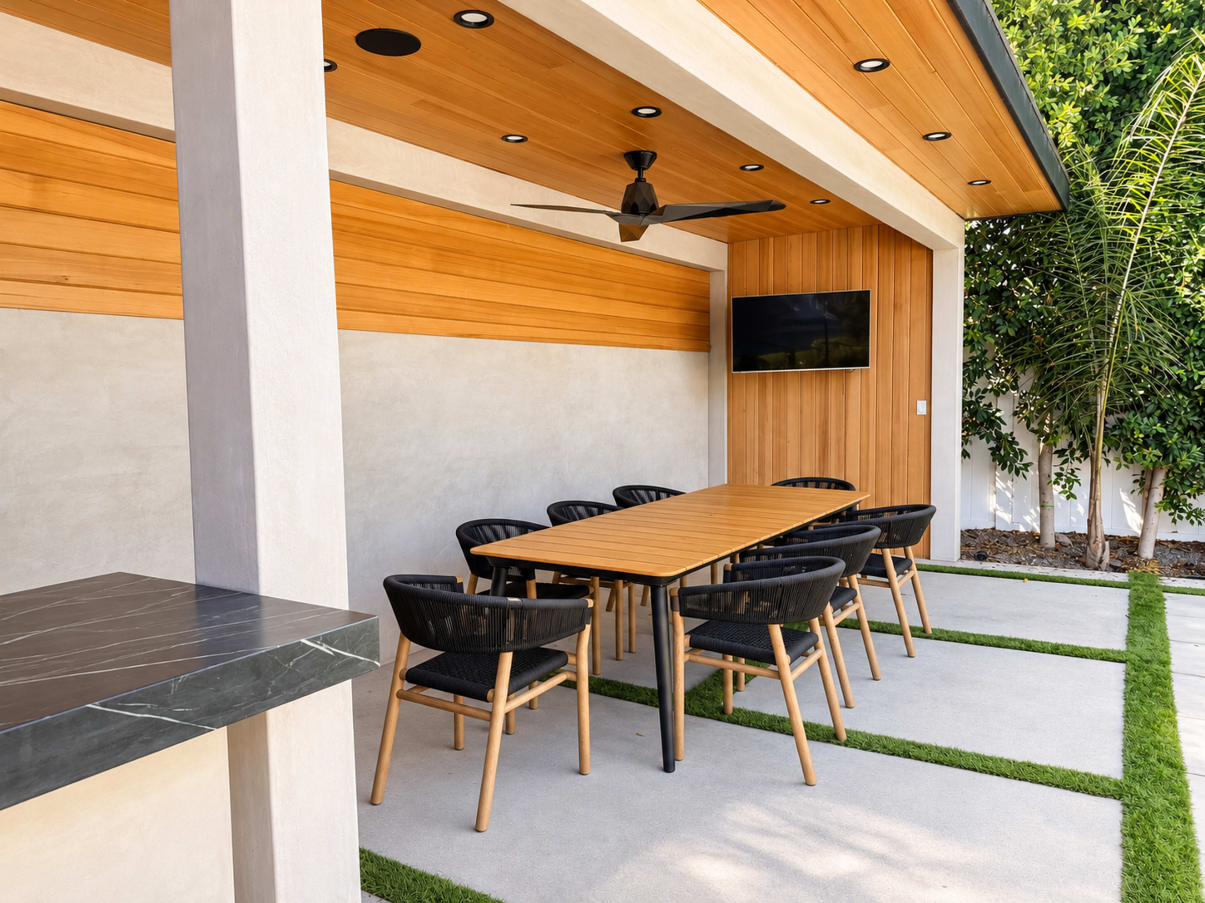 The image shows an outdoor dining area featuring a long wooden table surrounded by black chairs, with a ceiling fan and a wall-mounted TV in a modern architectural setting.
