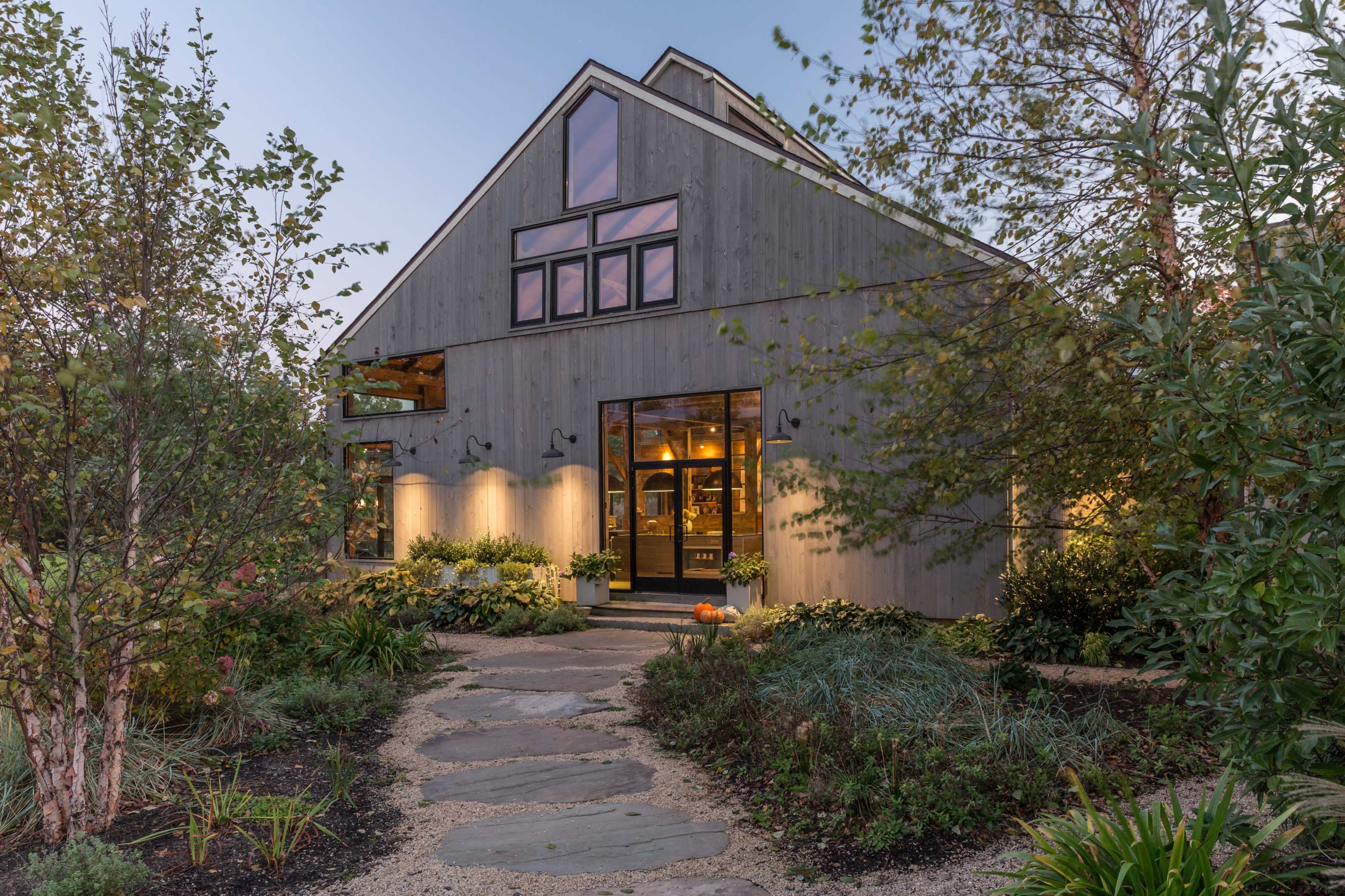 A modern gray barn-style house is surrounded by landscaped gardens and a stone pathway leading to its entrance.