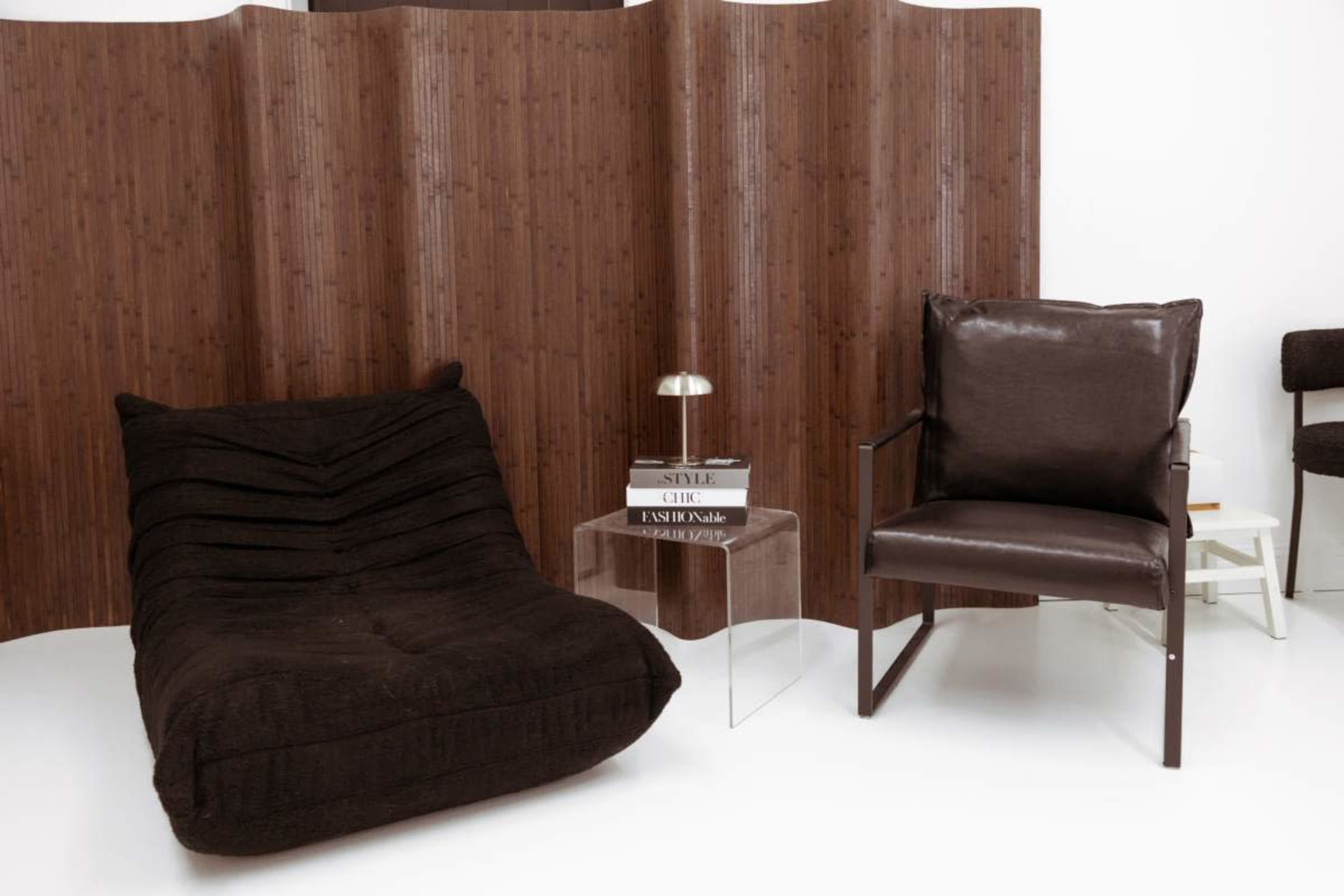 The image features a modern living space with a textured dark bean bag chair, a minimalist clear side table, and a sleek brown armchair set against a wooden room divider.
