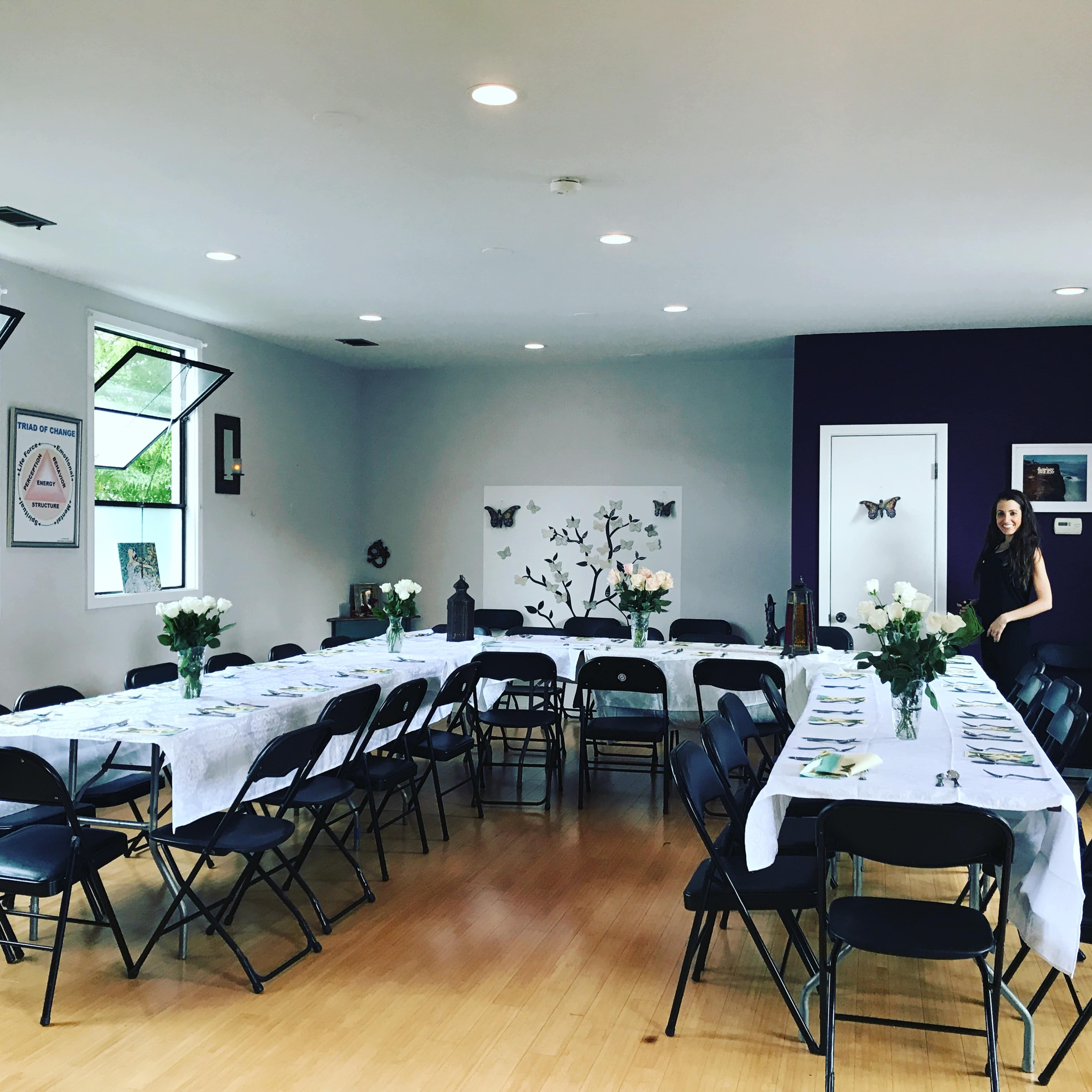 A neatly arranged dining area features multiple tables set with white tablecloths and centerpieces of white flowers, with a woman standing nearby against a purple wall.