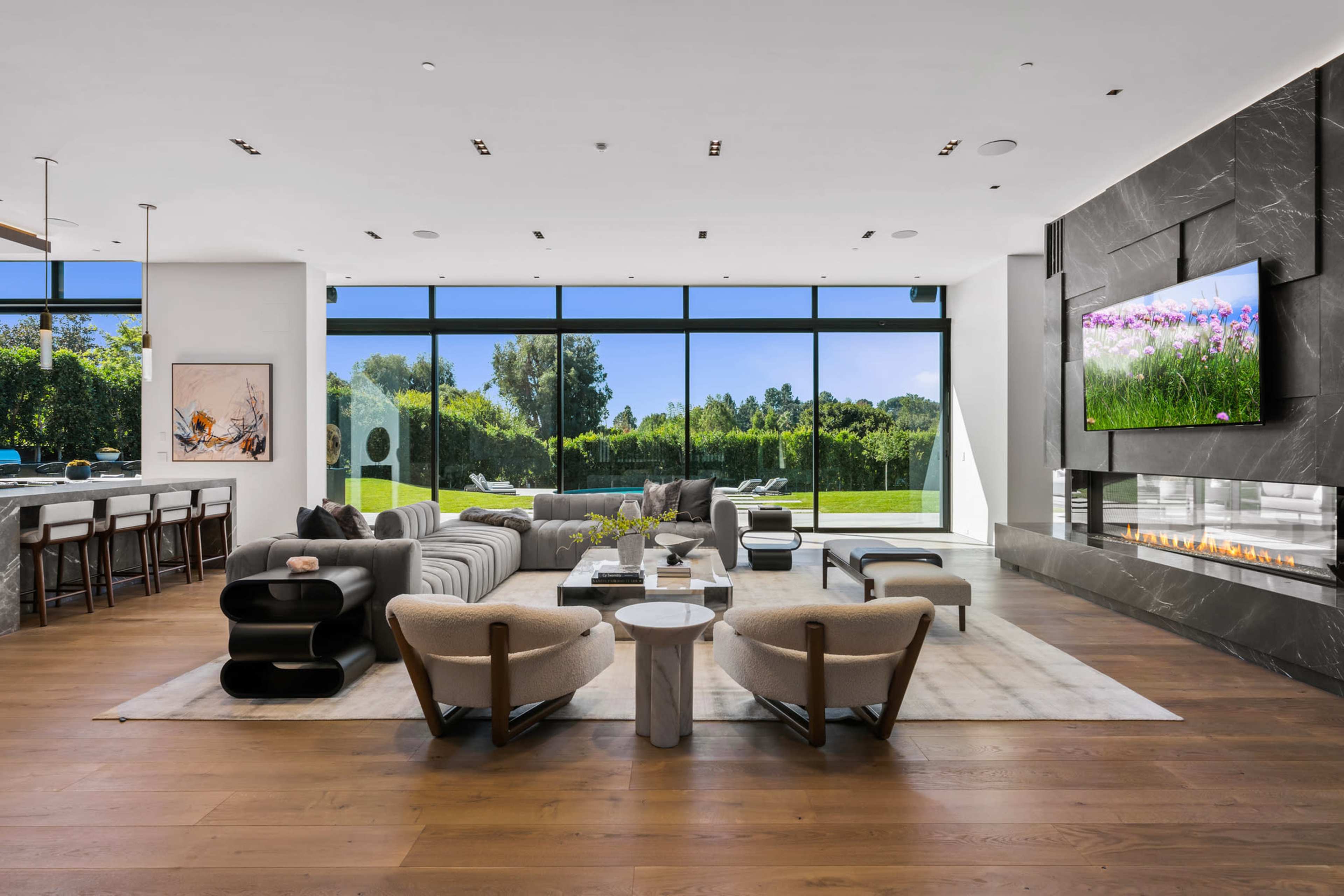 The image shows a modern living room with large windows displaying a green outdoor area, featuring a grey sectional sofa, armchairs, a coffee table, and a sleek fireplace.