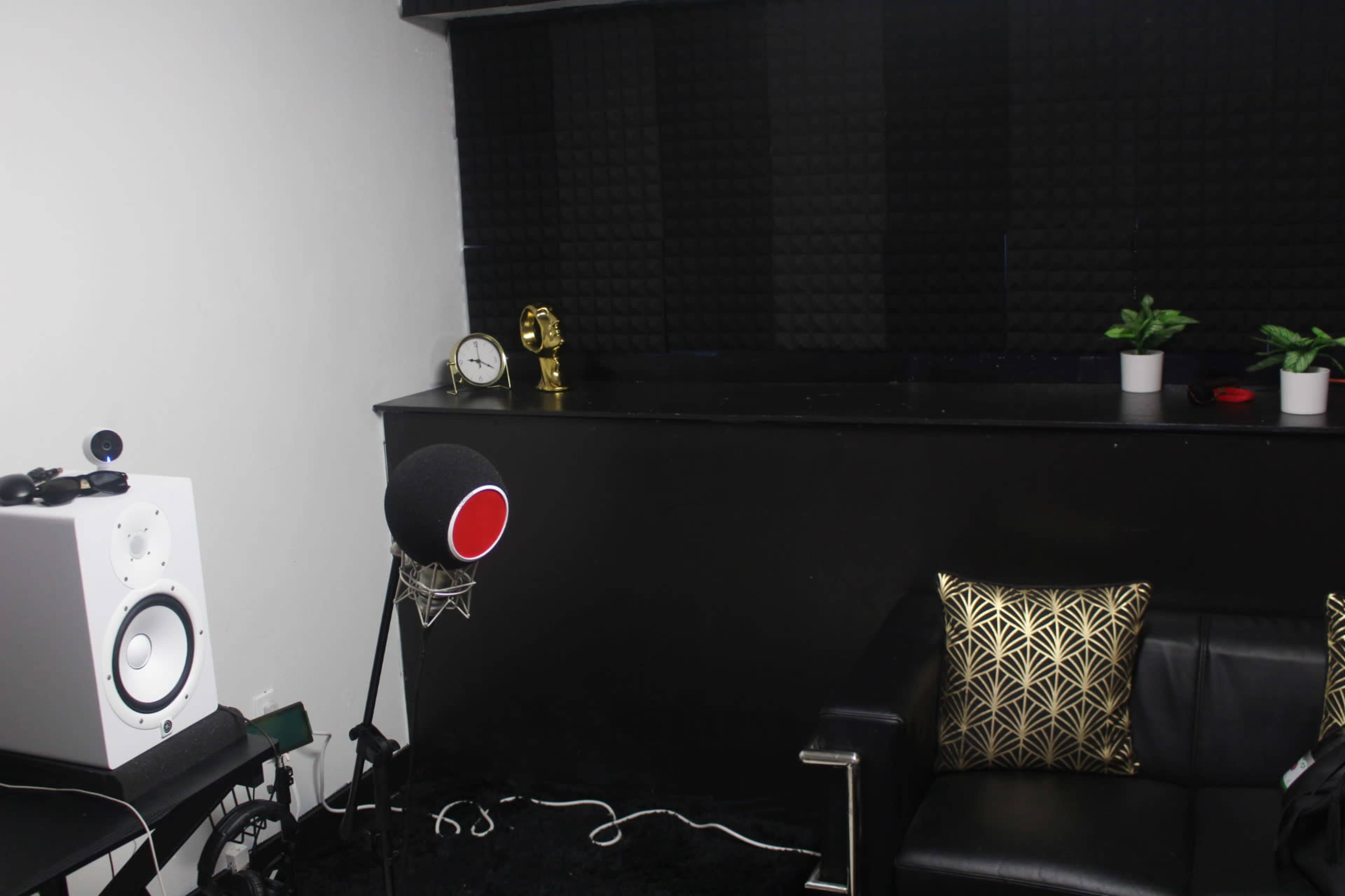 A recording studio corner features a black couch, a microphone with a pop filter, a speaker, a small clock, and decorative plants against a textured black wall.