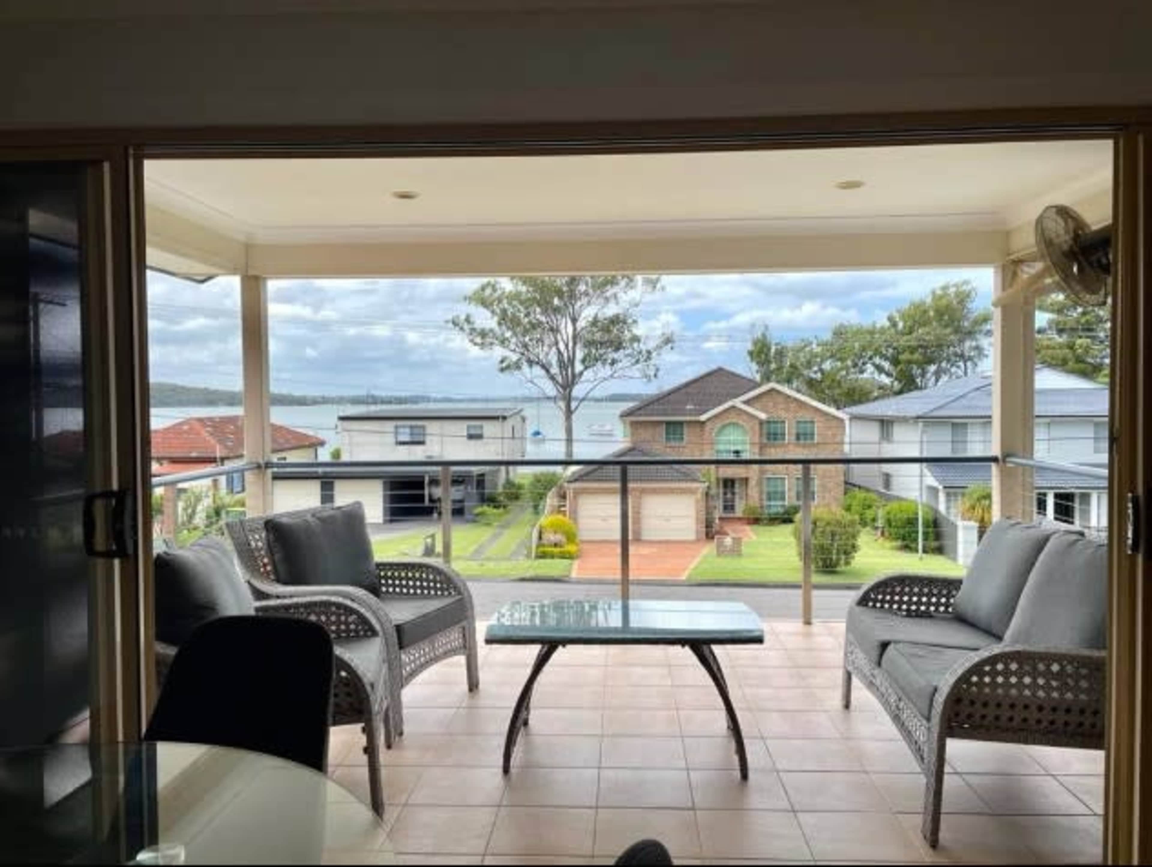 A balcony with two gray chairs and a glass table, overlooking a landscaped yard and houses adjacent to a body of water.