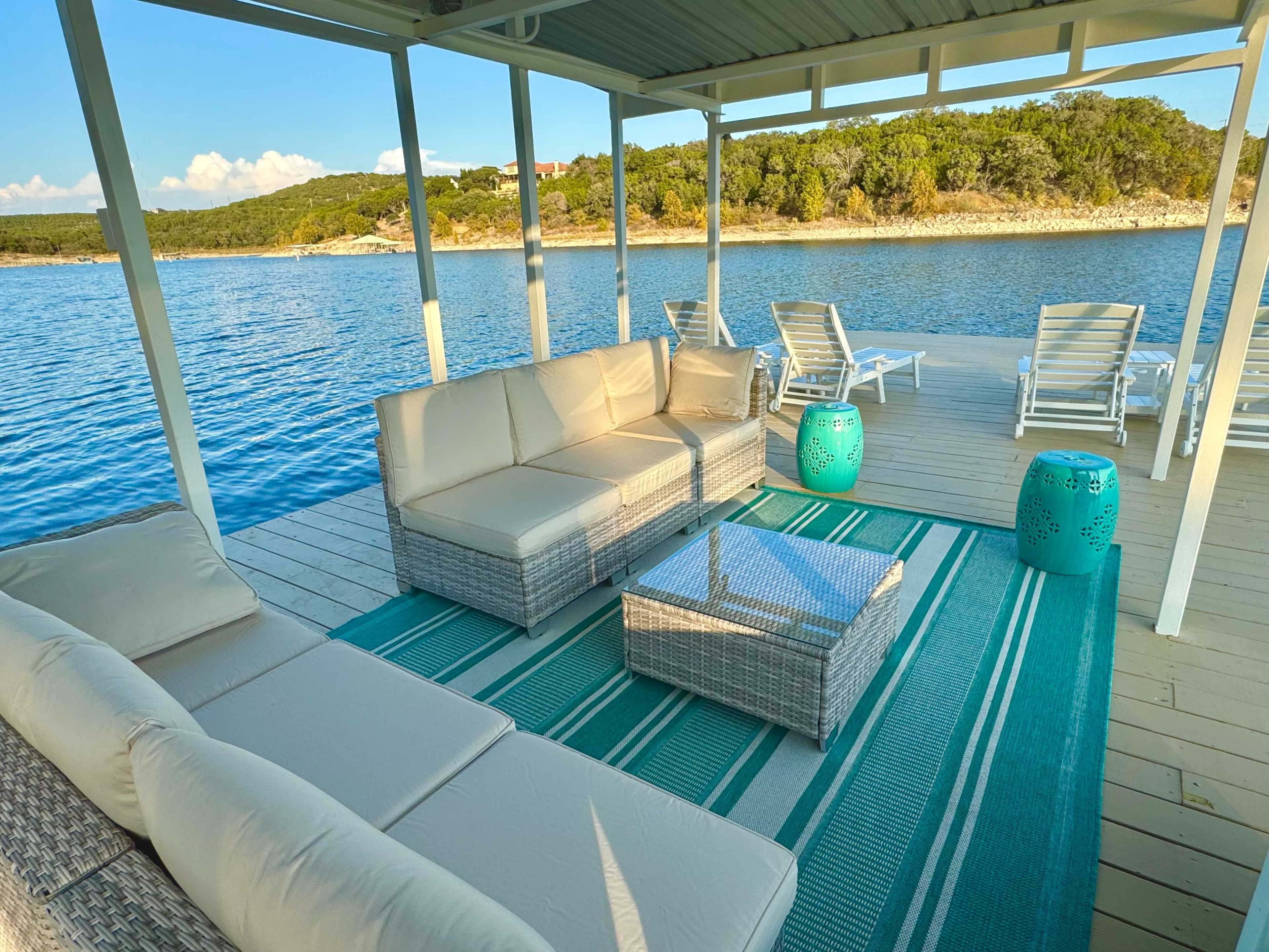 The image shows a comfortable seating area on a dock by a calm lake, featuring a sectional sofa, a glass coffee table, and turquoise accent tables overlooking the water.