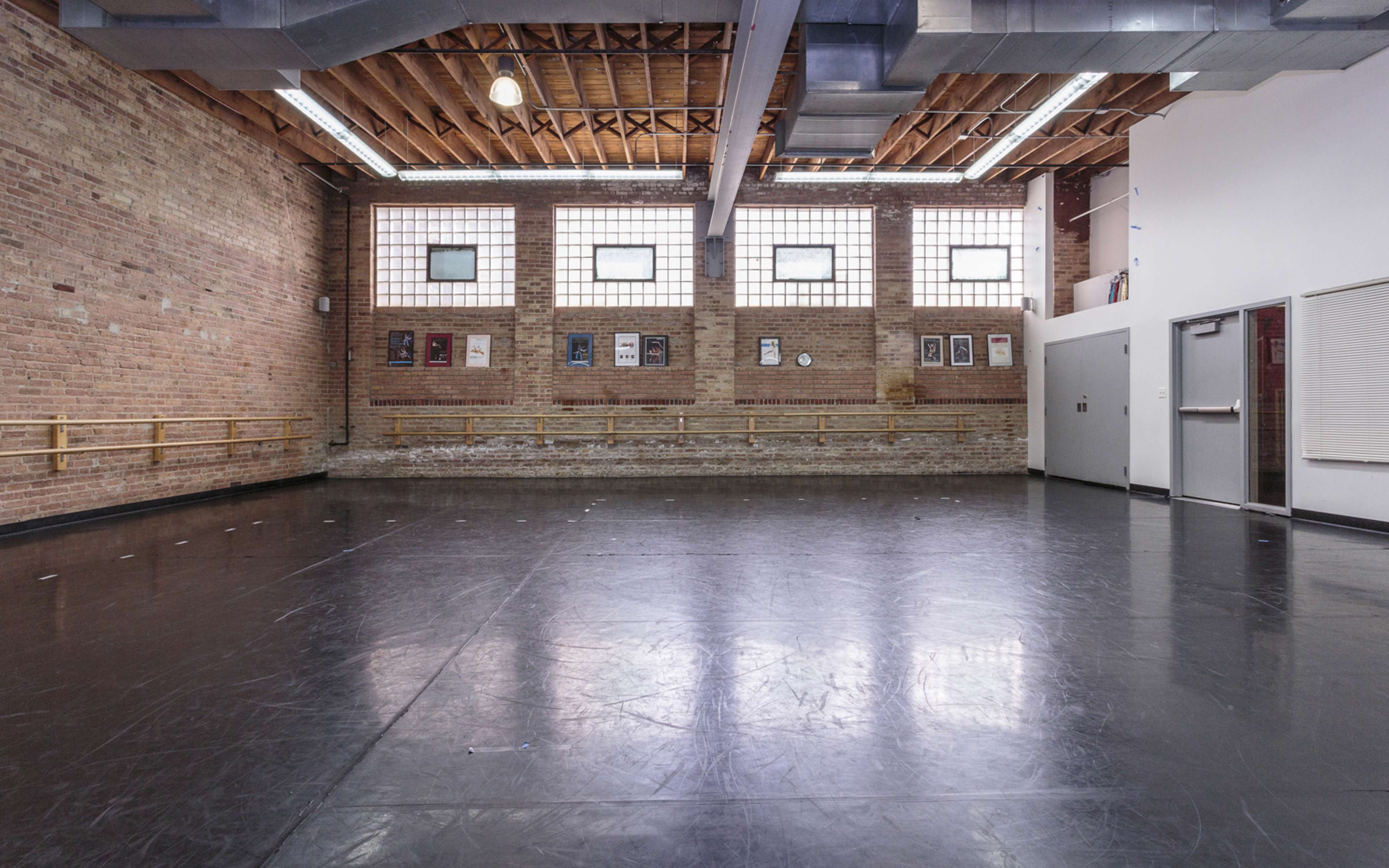 An empty dance studio with hardwood floors, exposed brick walls, large windows, and ballet barres along the walls.