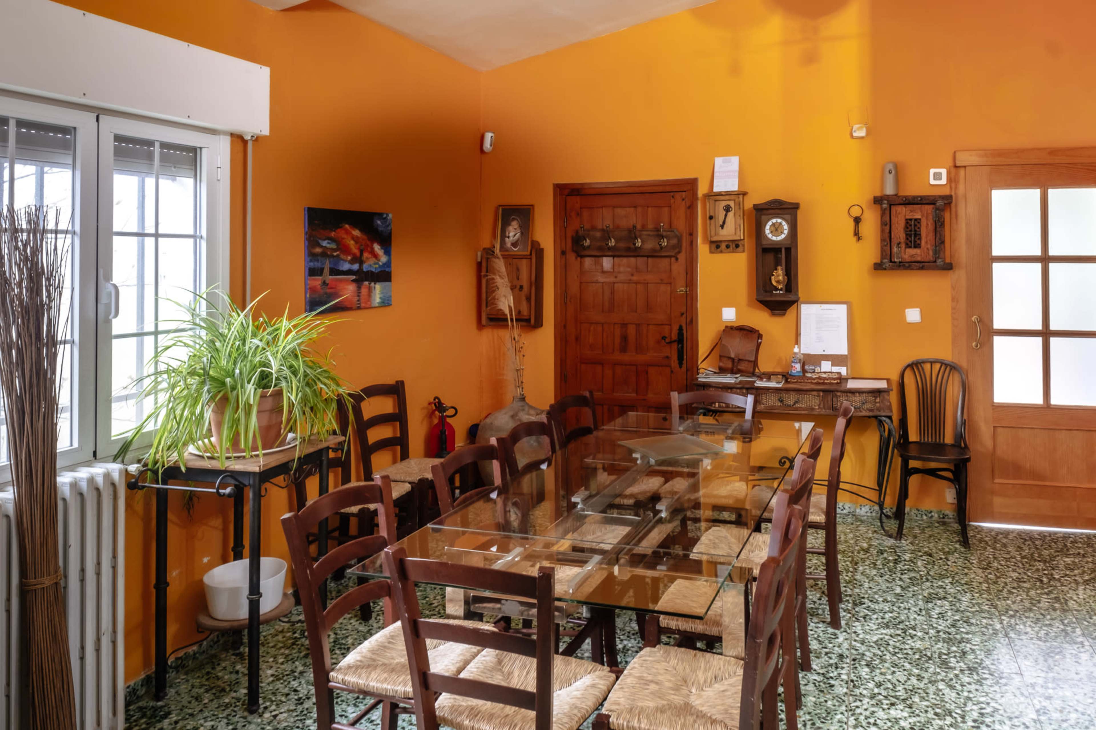 A dining area features a large glass table surrounded by wooden chairs against an orange wall, with various decorations and a window letting in natural light.