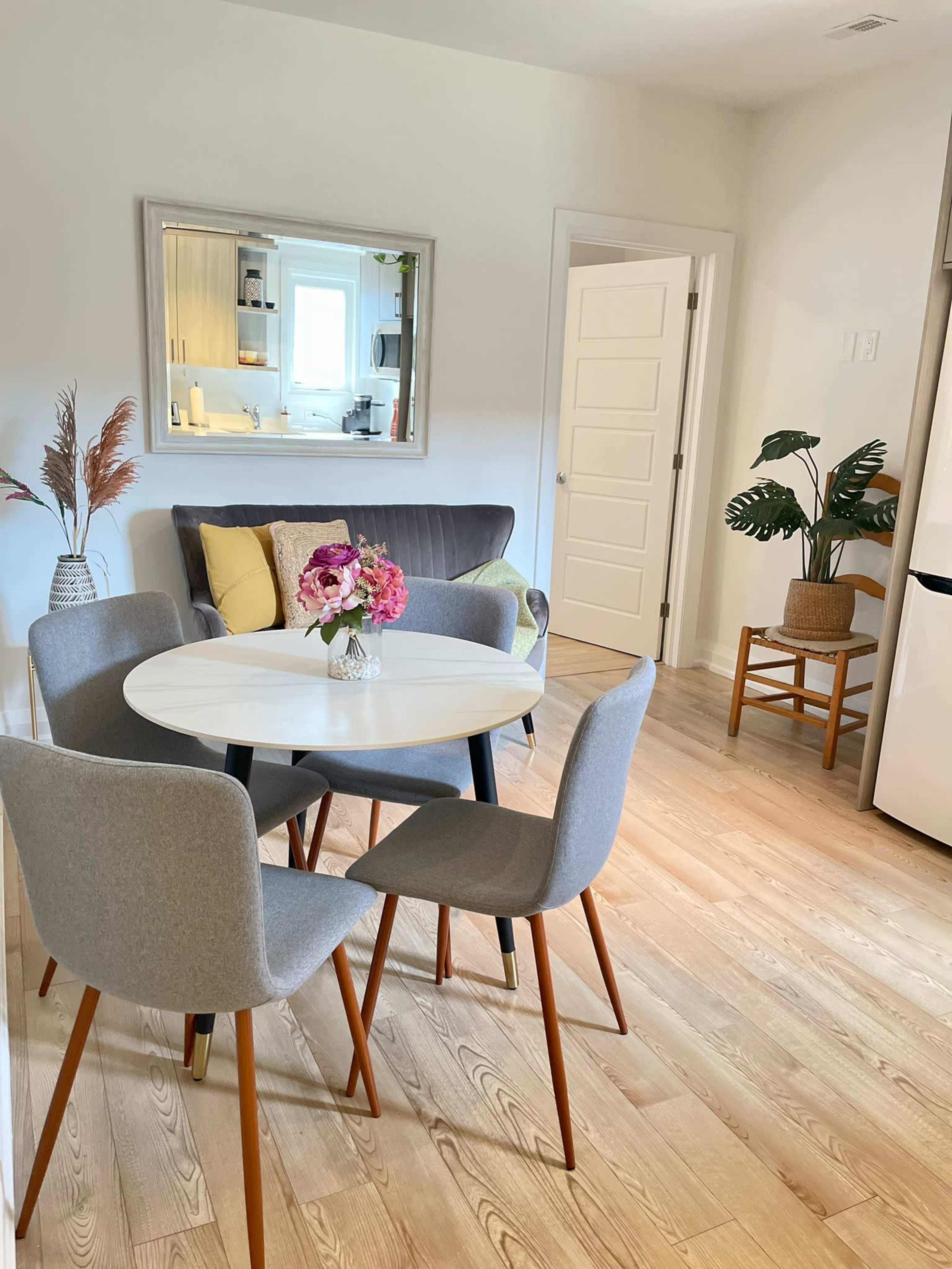 A dining area features a round table with gray chairs, a small vase of flowers, a gray sofa, and a mirror on the wall.