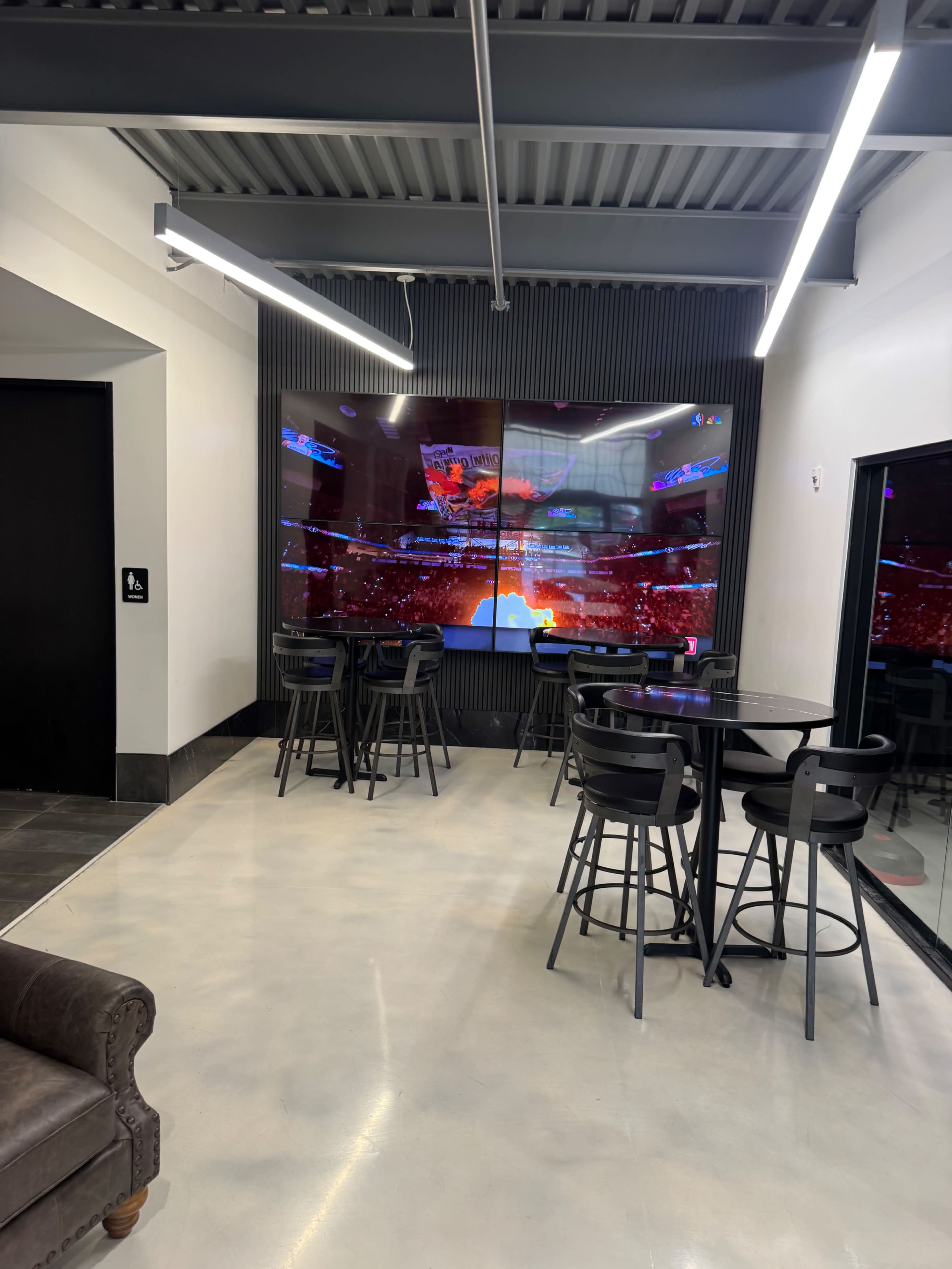The image shows a modern lounge area with black bar stools and tables, featuring a large screen displaying a vibrant event in the background.