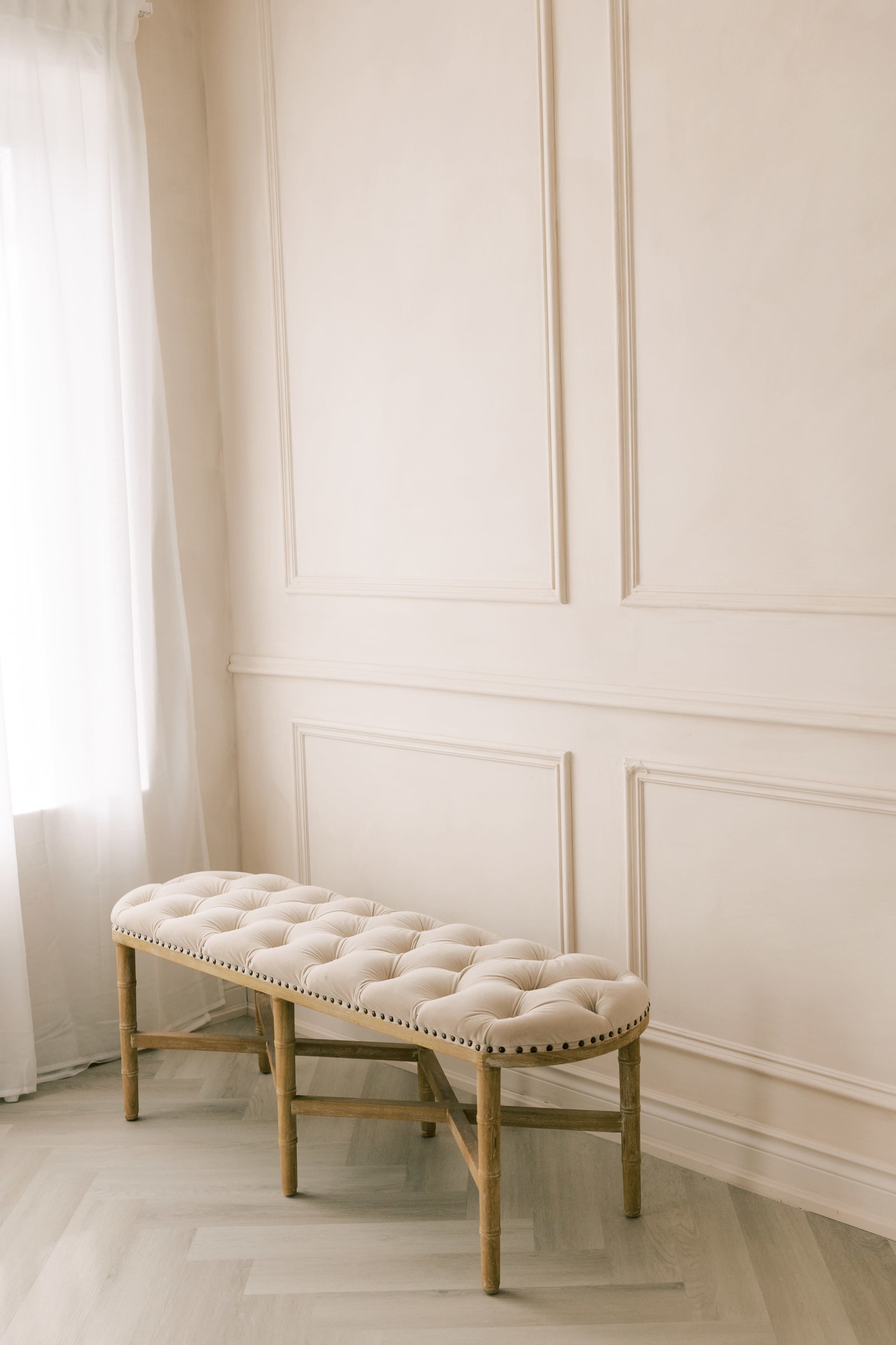 A tufted beige bench sits against a light-colored wall in a brightly lit room.