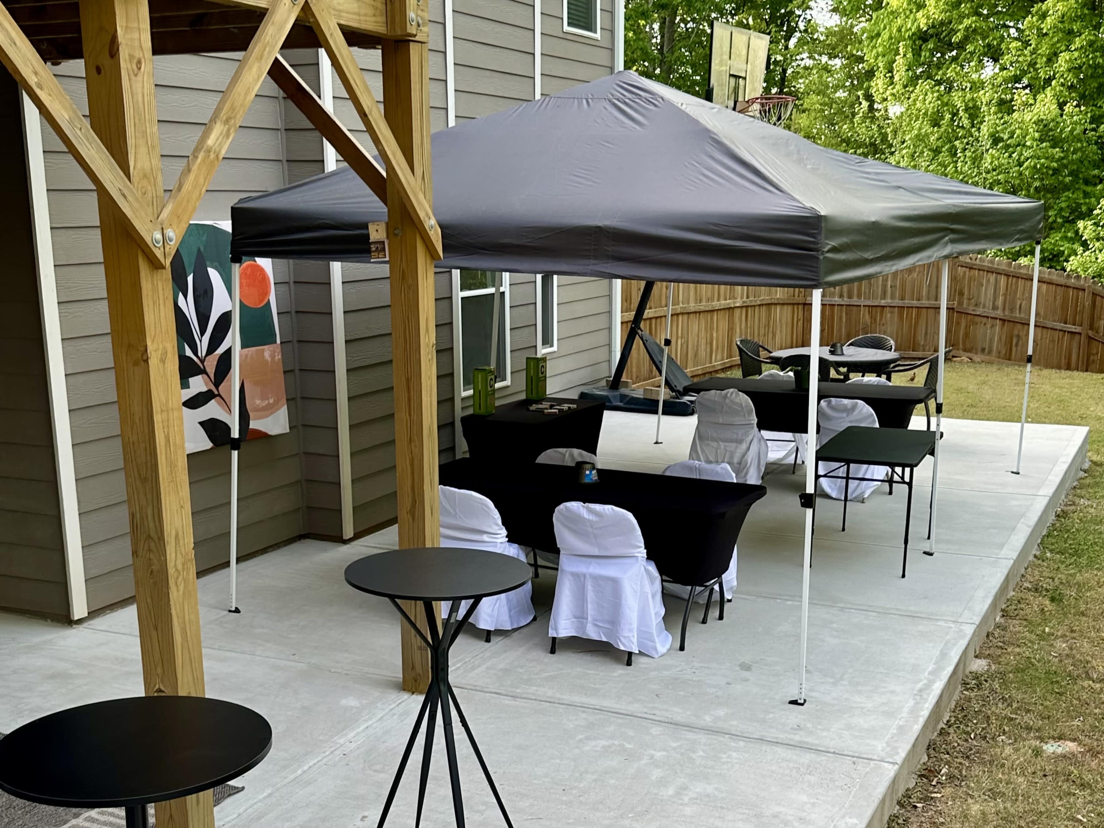 A shaded outdoor seating area features a gray canopy, black chairs, and small tables near a house with a basketball hoop.