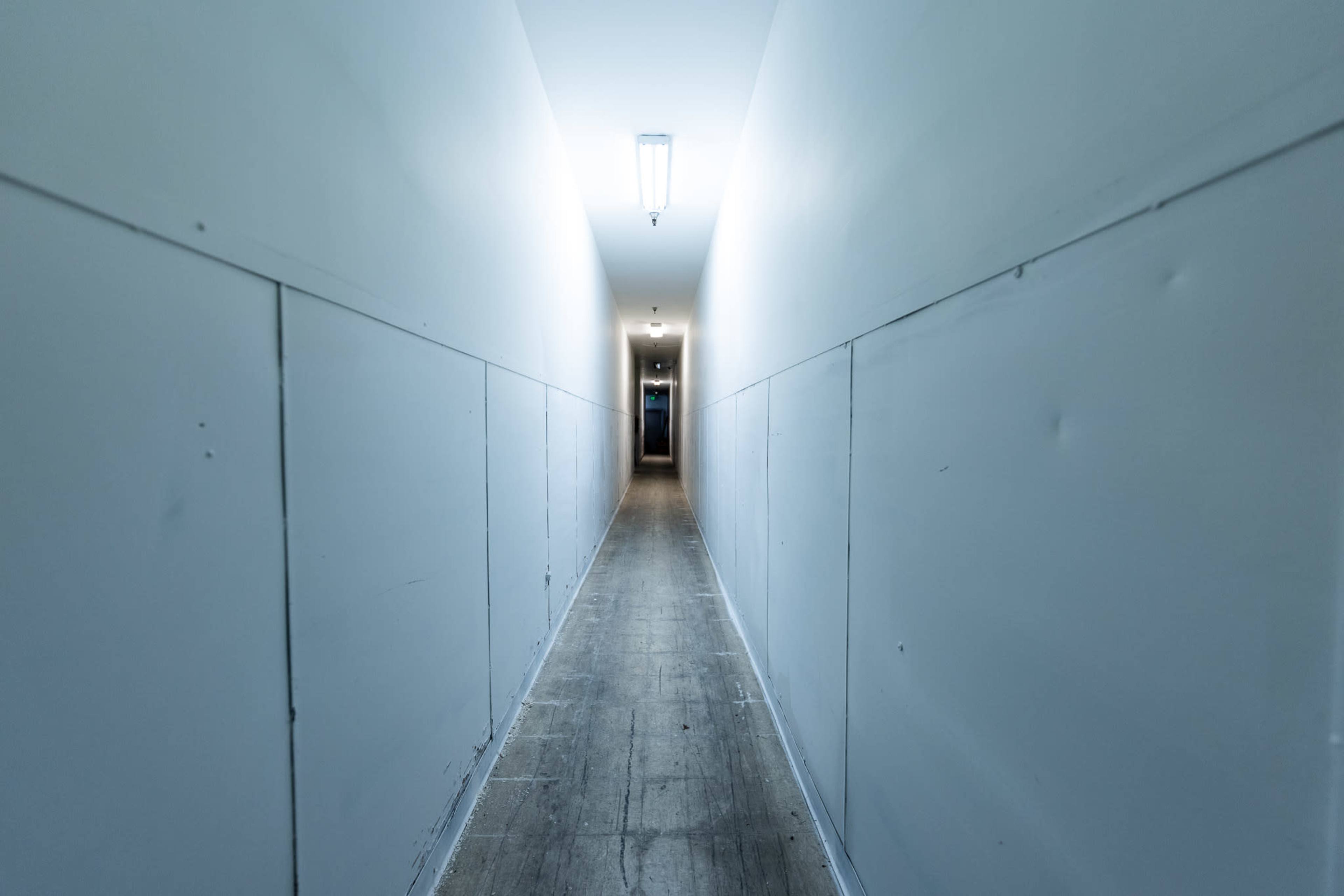 A long, narrow hallway with white walls and a light fixture at the ceiling leads to a distant doorway.