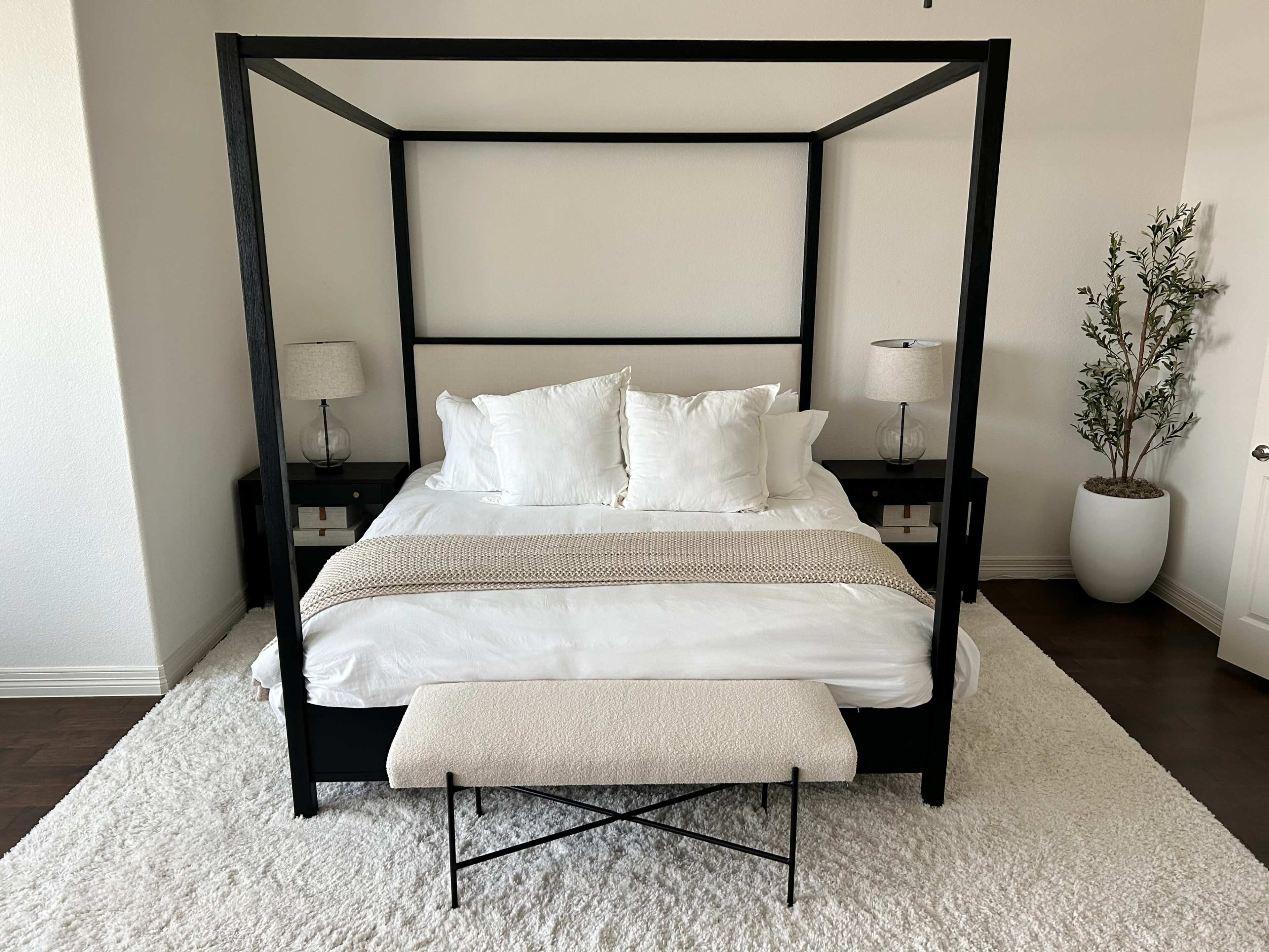A black four-poster bed with white bedding and two bedside lamps is centered on a plush white rug in a well-lit room.
