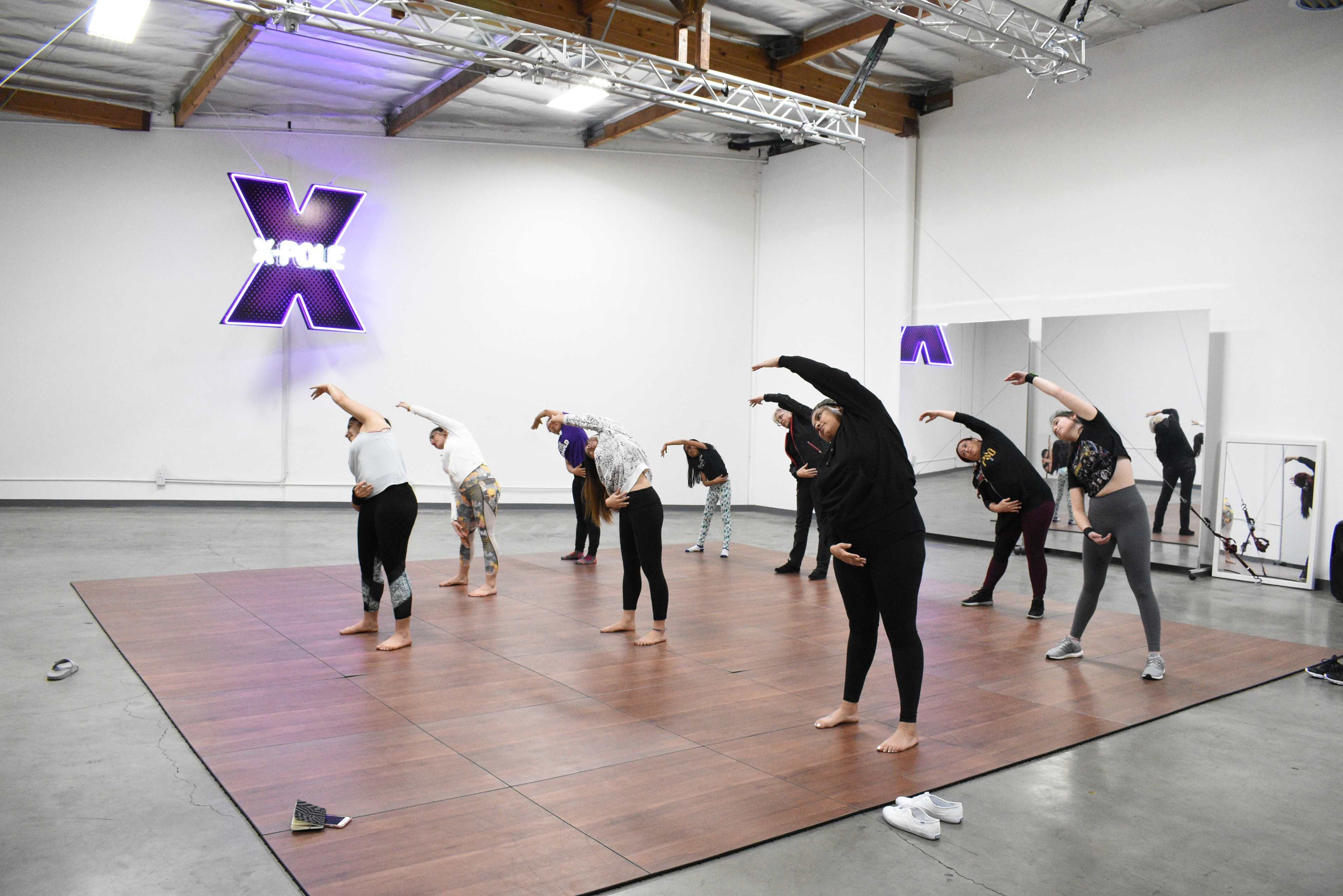 A group of people is practicing stretching exercises on a wooden dance floor in a studio with neon lighting.