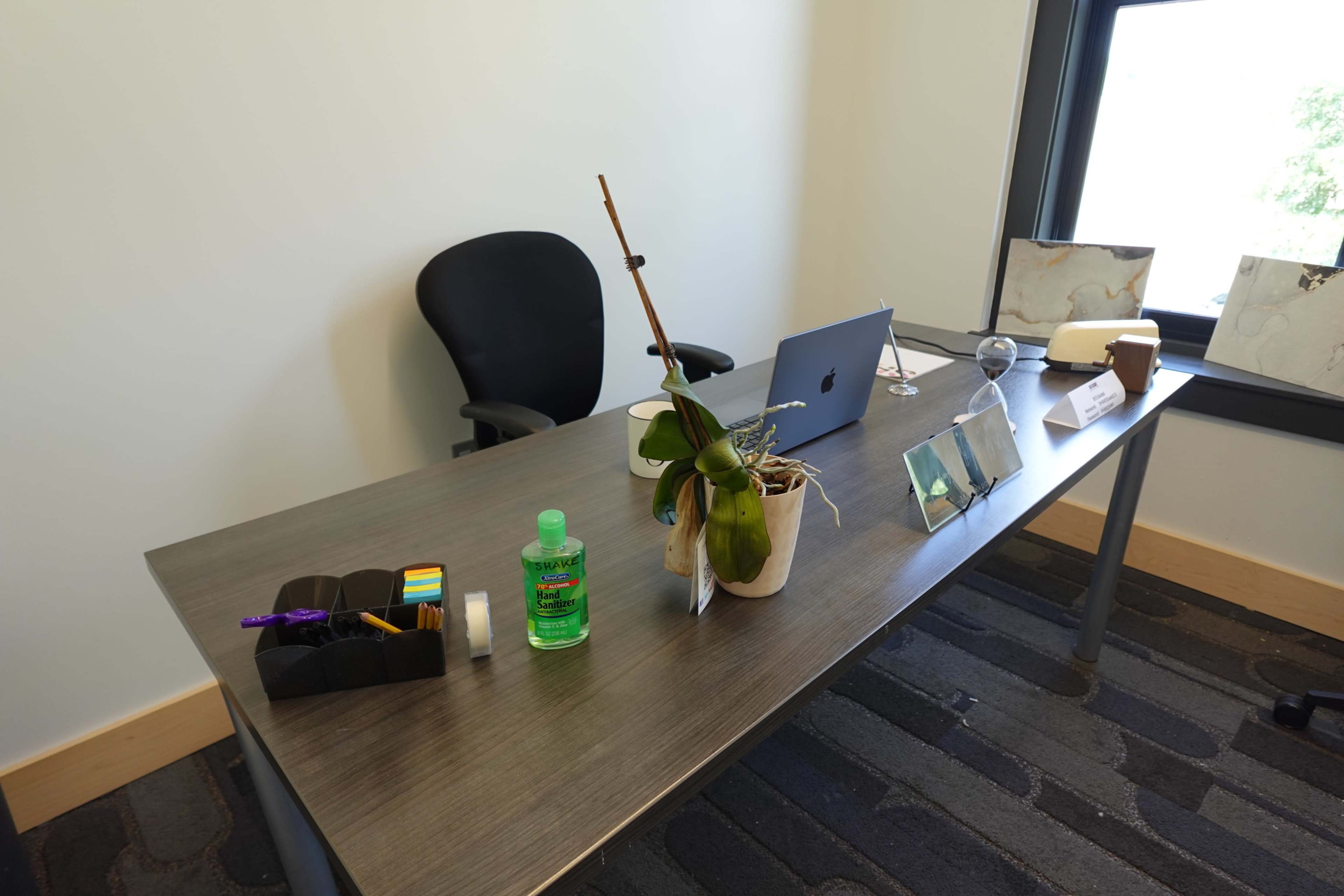 The image shows a neatly organized office desk with a computer, office supplies, a plant, and a chair in a brightly lit room.