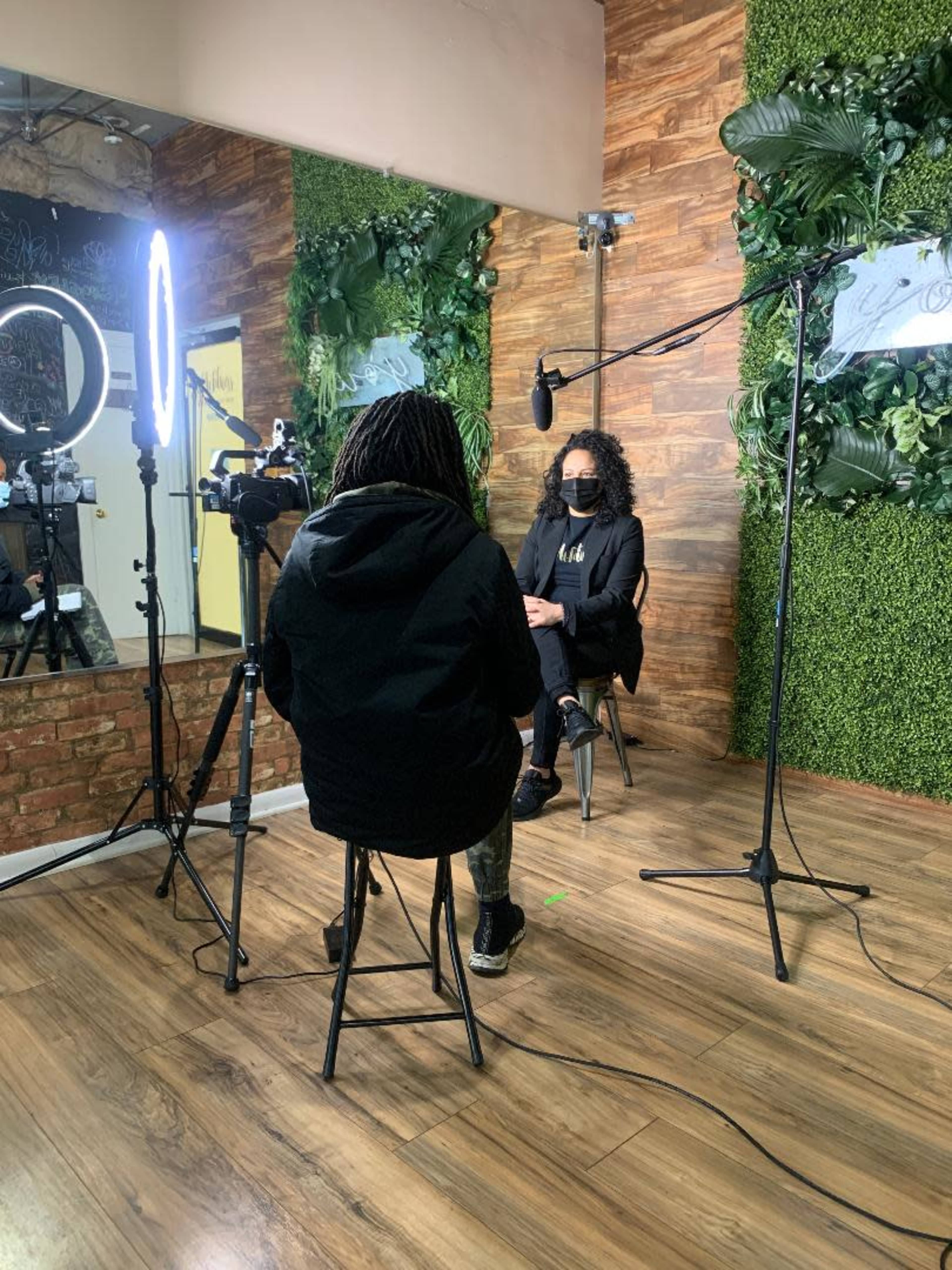 A person wearing a black hoodie sits on a chair facing a woman in a black outfit, both positioned in a studio environment with greenery and lights.