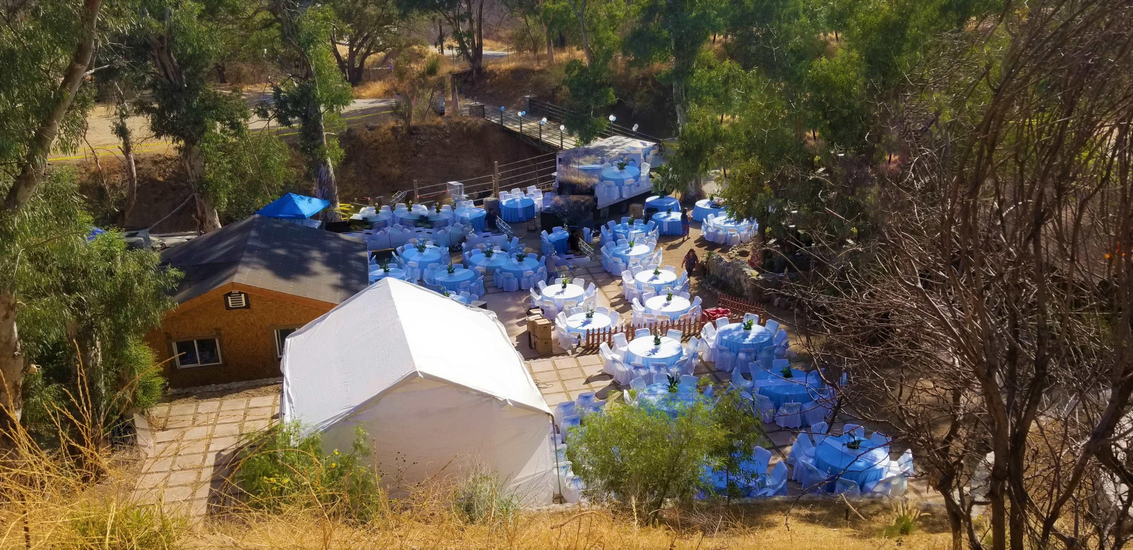 An outdoor event setup with numerous round tables covered in light blue tablecloths surrounded by greenery.