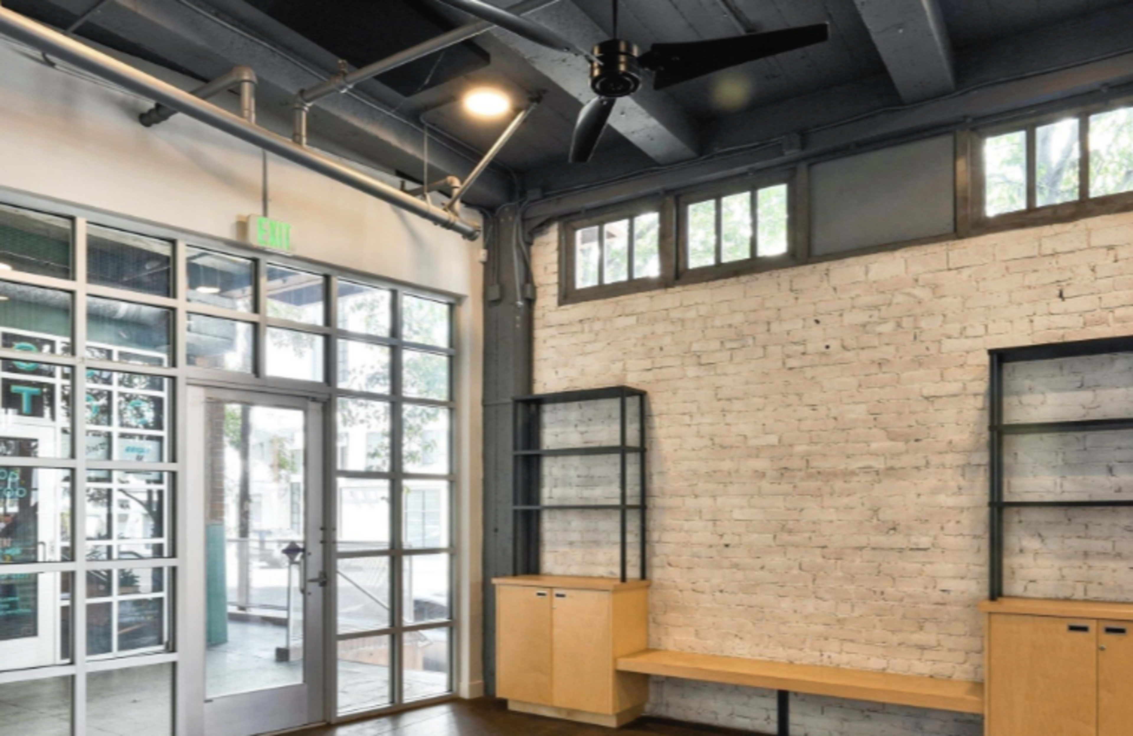 The image features a modern interior space with a glass entrance, exposed brick walls, wooden shelves, and a ceiling fan.