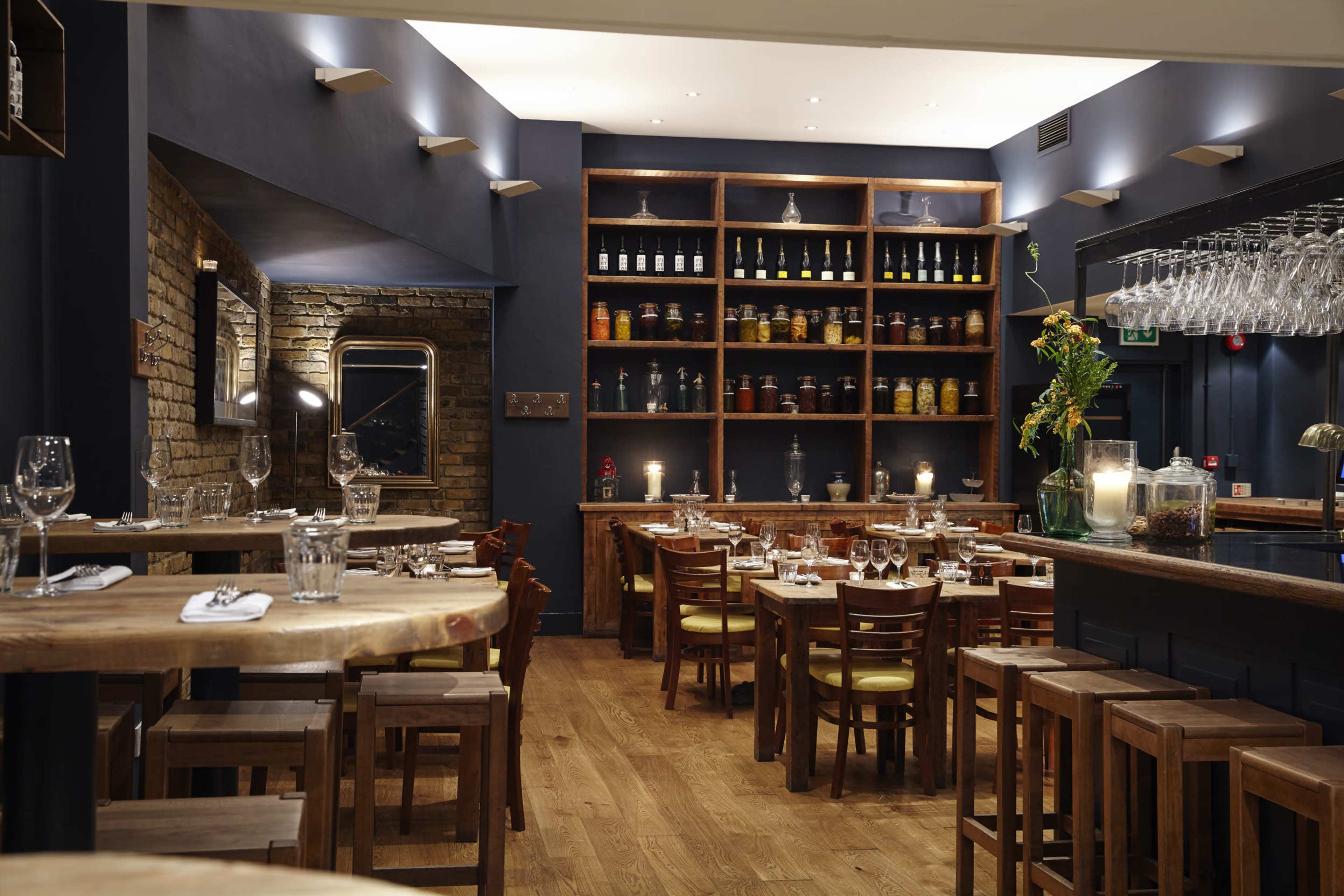 The image depicts a restaurant interior featuring wooden tables and chairs, a wall-lined shelf with jars, and ambient lighting.